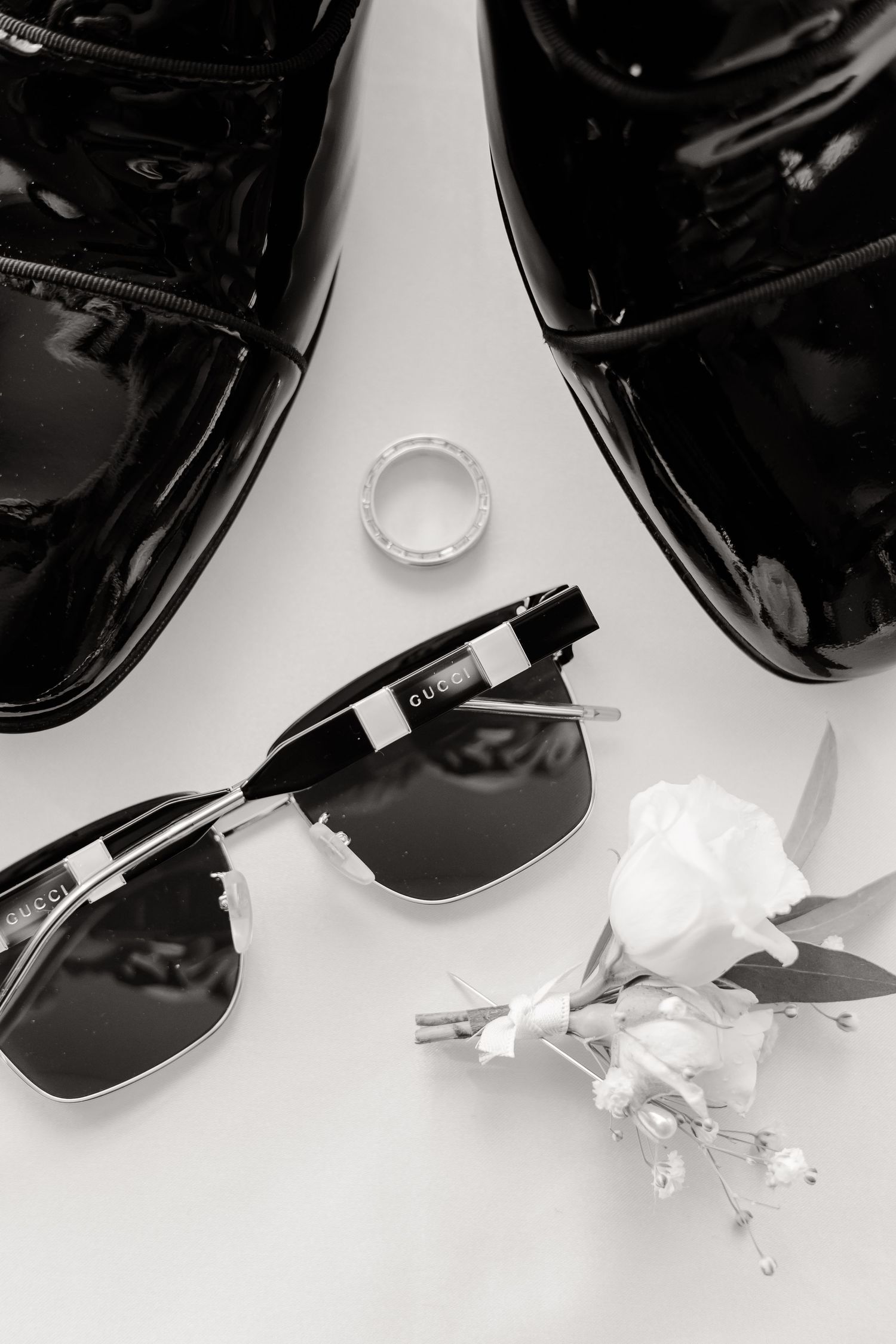 Black and white photo of dress shoes with sunglasses and wedding ring on white surface.