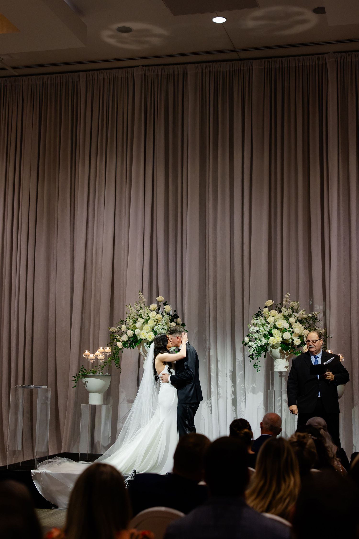 Wedding ceremony captured from audience perspective showing candlelit floral decor and draped backdrop.