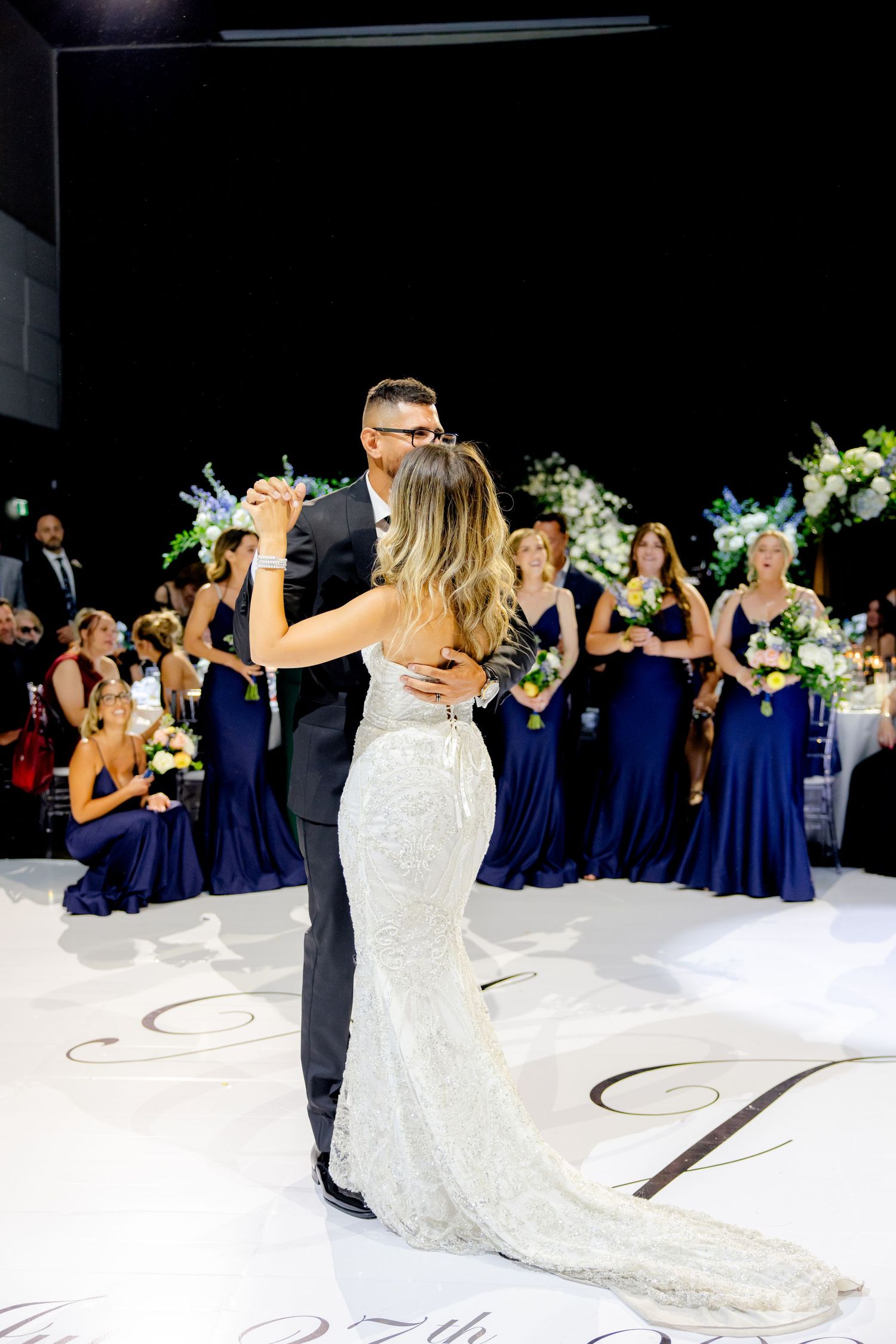 A wedding couple shares an intimate dance on a white dance floor surrounded by their wedding party.