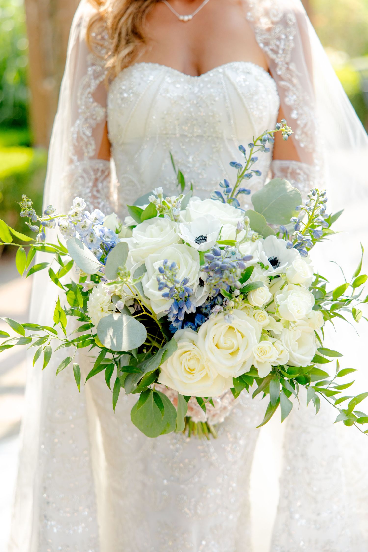 Close-up of white and blue bridal bouquet with roses, delphinium and eucalyptus against lace wedding dress.
