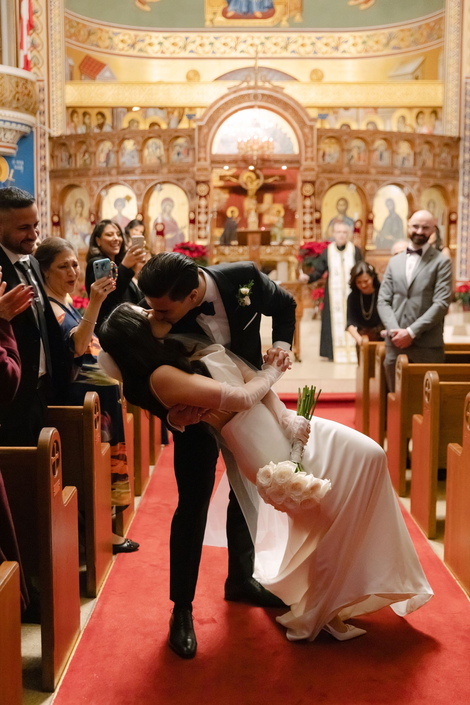A couple shares a romantic dip kiss in a grand church with ornate architecture and guests capturing the moment on phones.