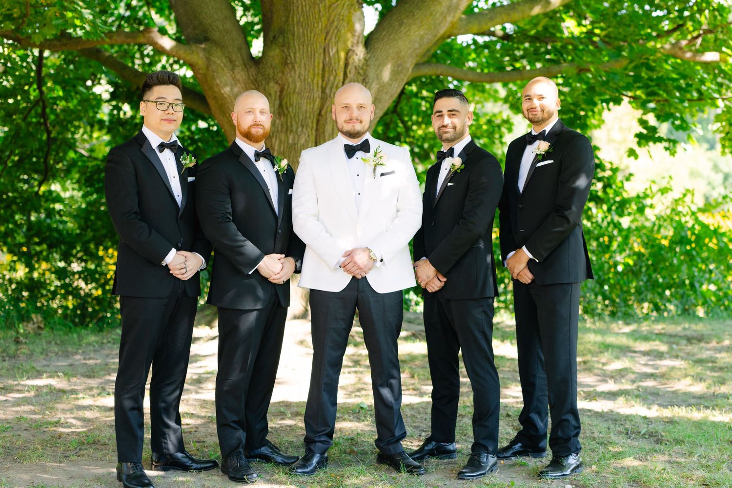 Groomsmen in black tuxedos pose with groom in white jacket under green trees.