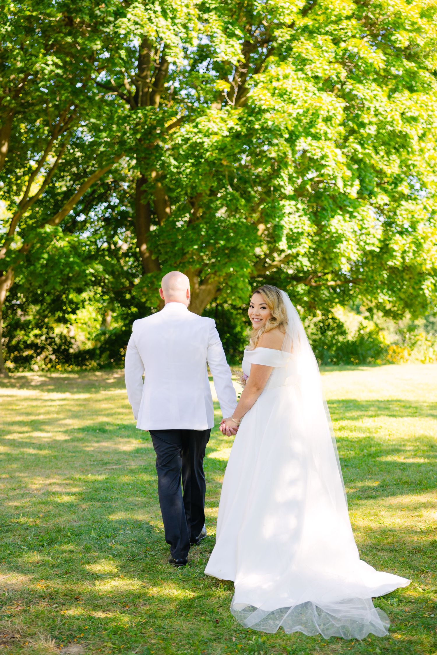 Newlyweds walk away from camera on grass path under lush green trees.