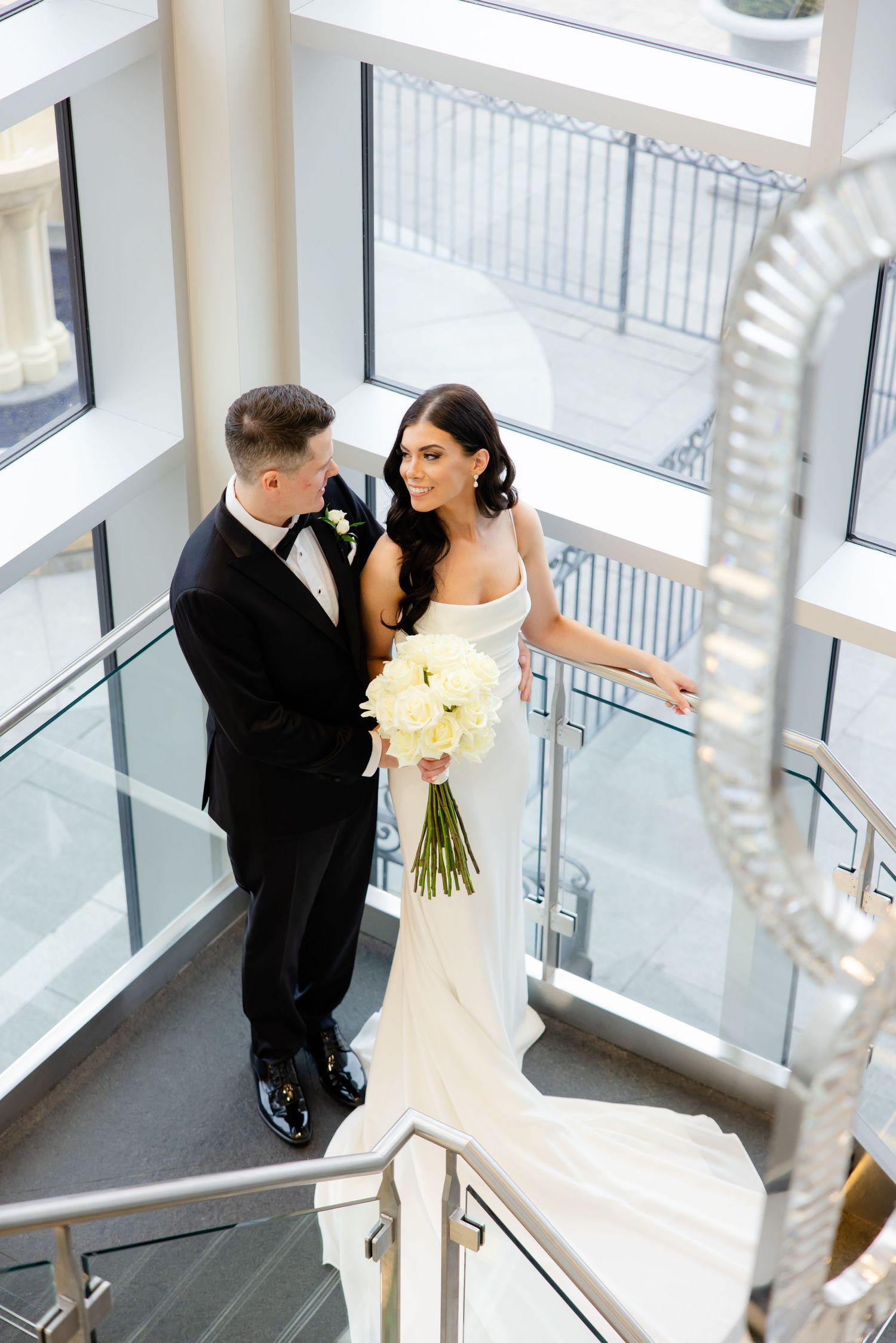 Modern wedding portrait on architectural staircase with hanging light fixtures and glass walls.