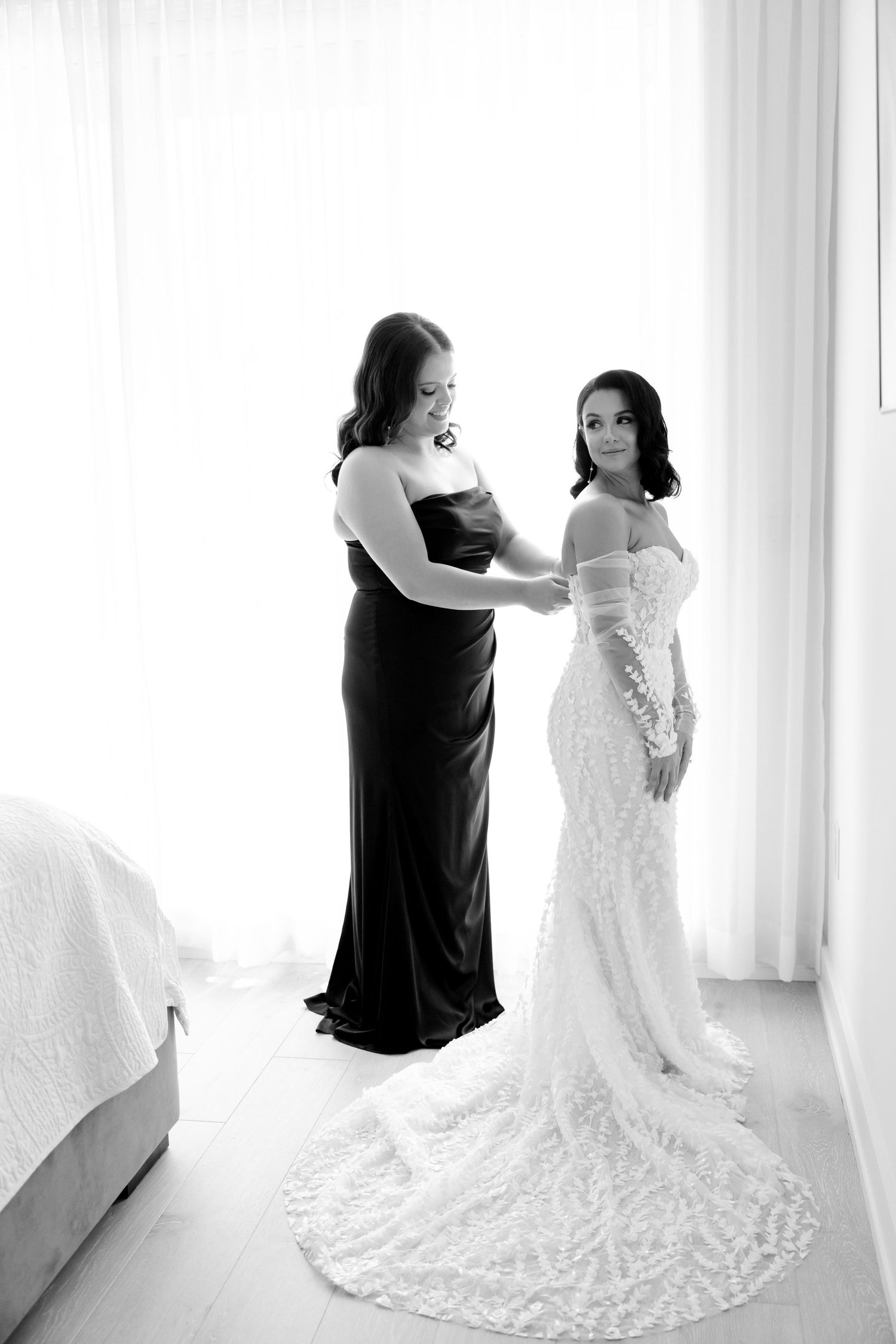 A bride in a lace wedding gown with a long train stands with her bridesmaid in a black dress by a window.