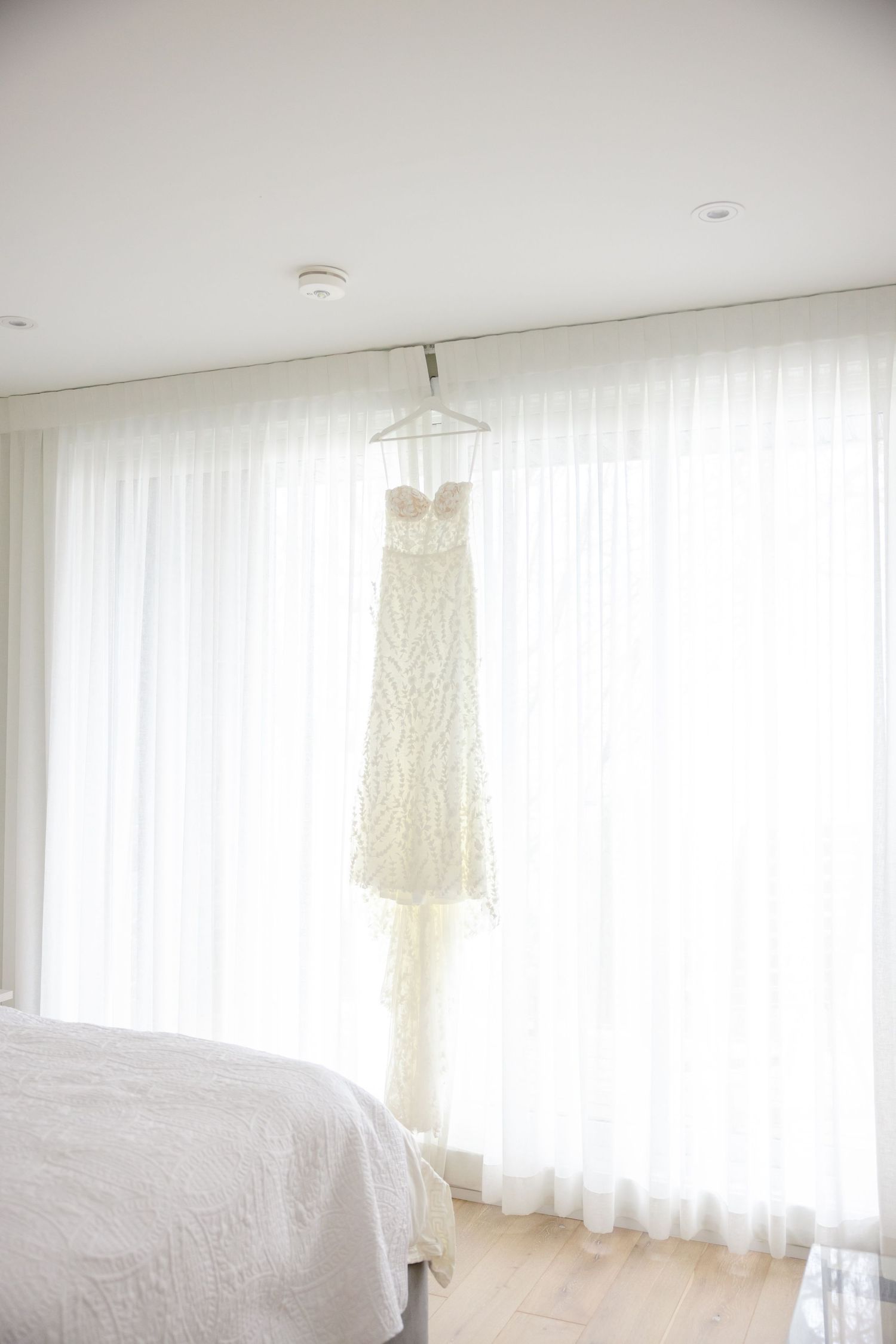 White lace wedding dress hanging against sheer curtains in bright bedroom.