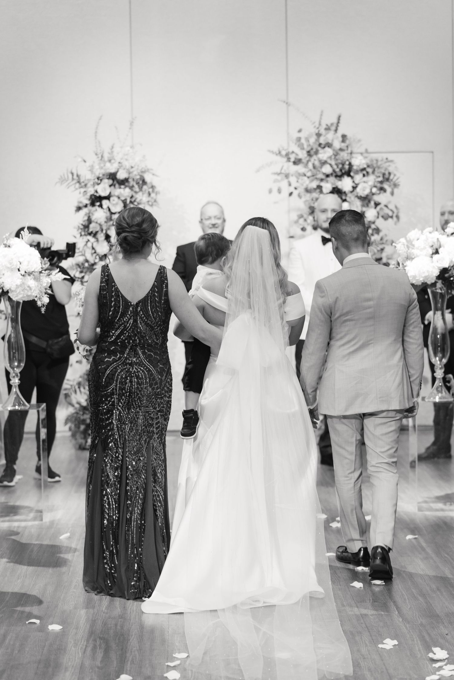 A black and white wedding ceremony photo showing guests and a bride from behind at the altar.