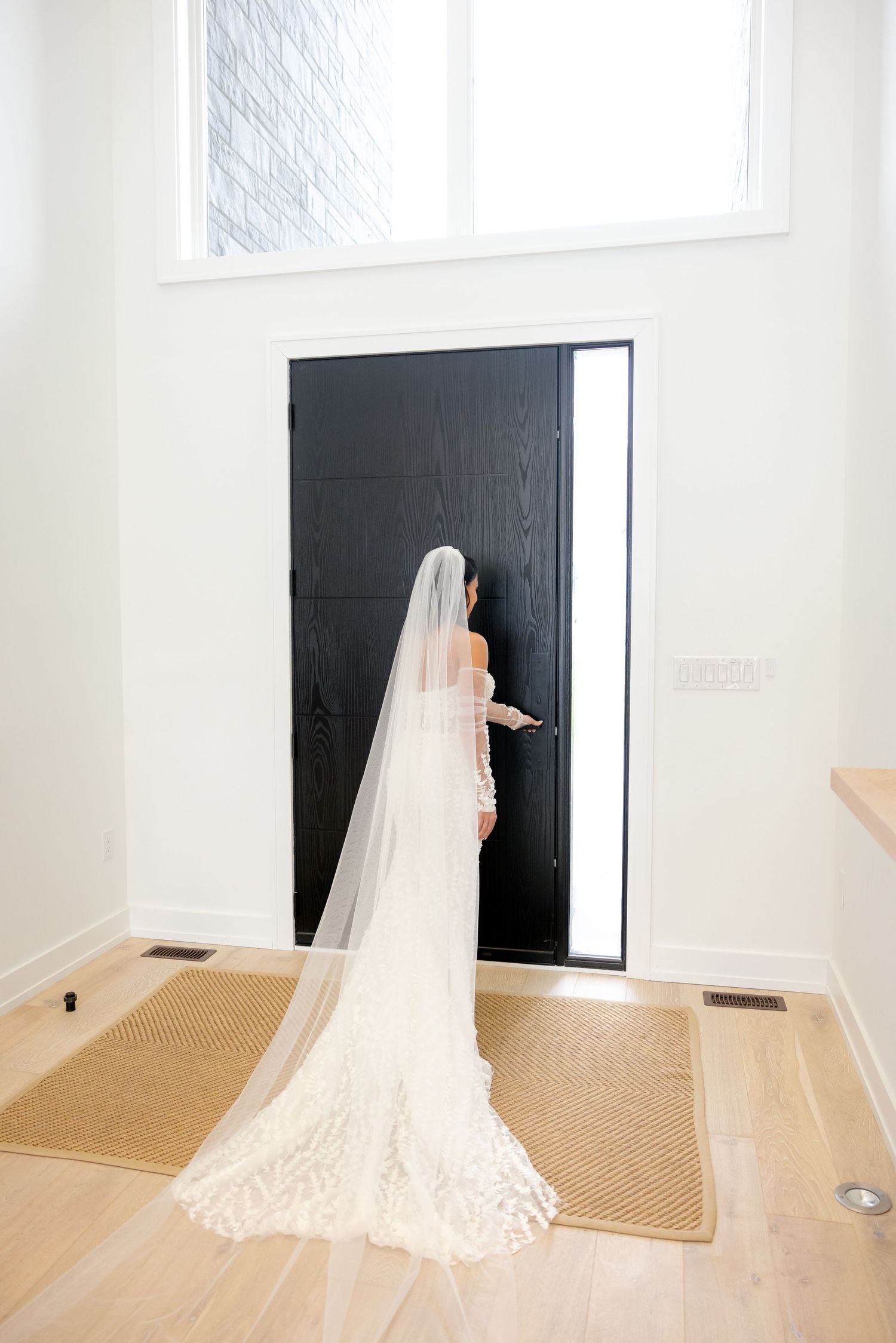 A bride in a long veil approaches a modern black door in her wedding dress.