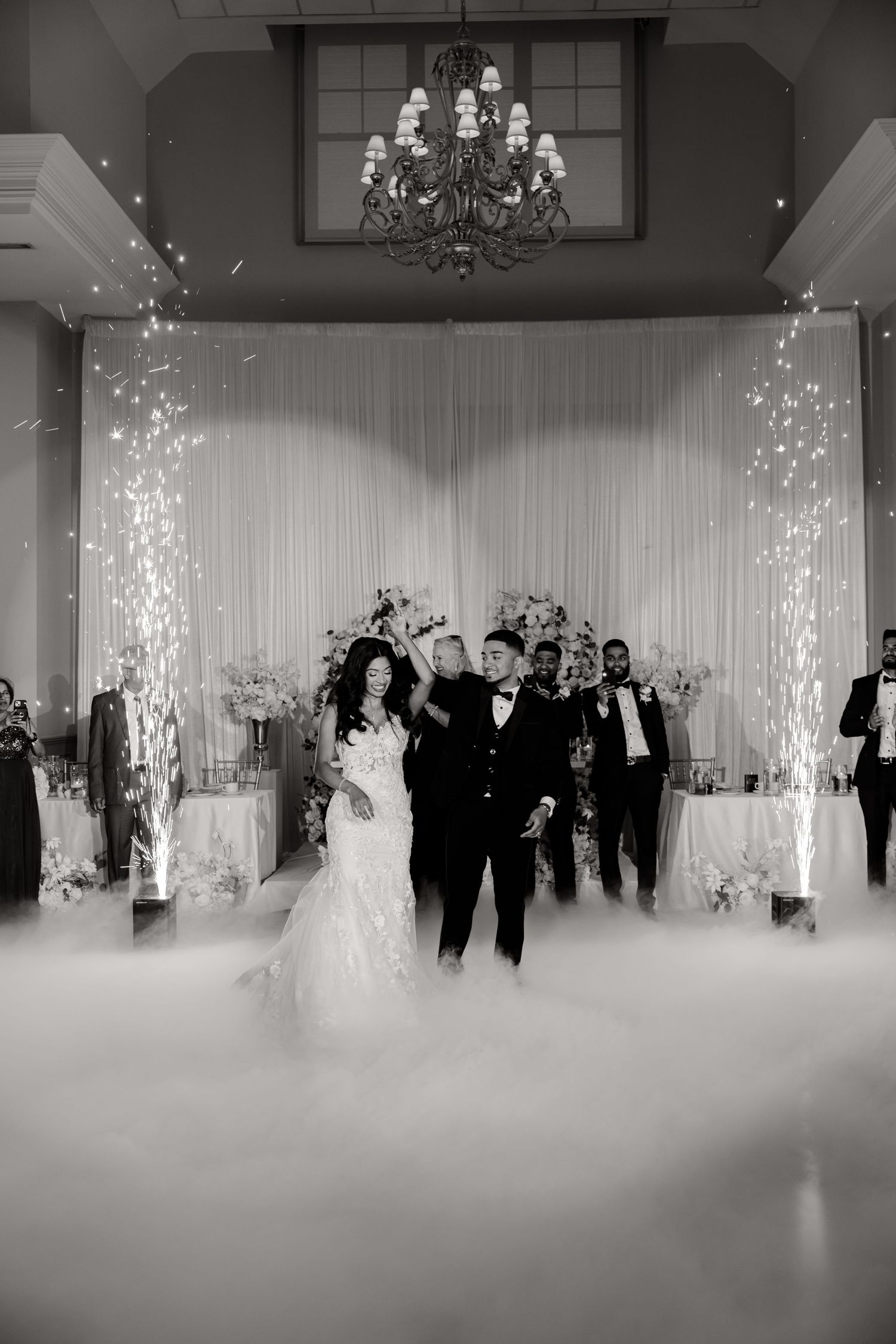 Elegant wedding ceremony entrance with sparklers and fog effects under a grand chandelier in black and white.