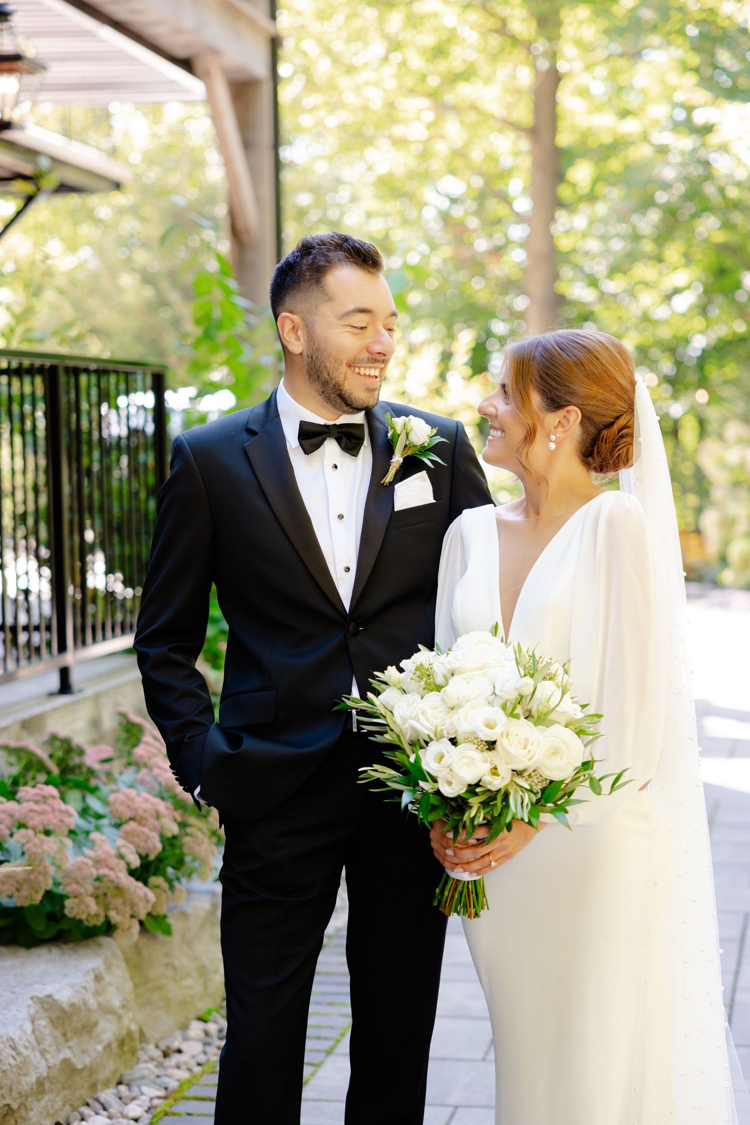 A joyful wedding couple in formal attire share a laugh while holding a white floral bouquet on a garden path.