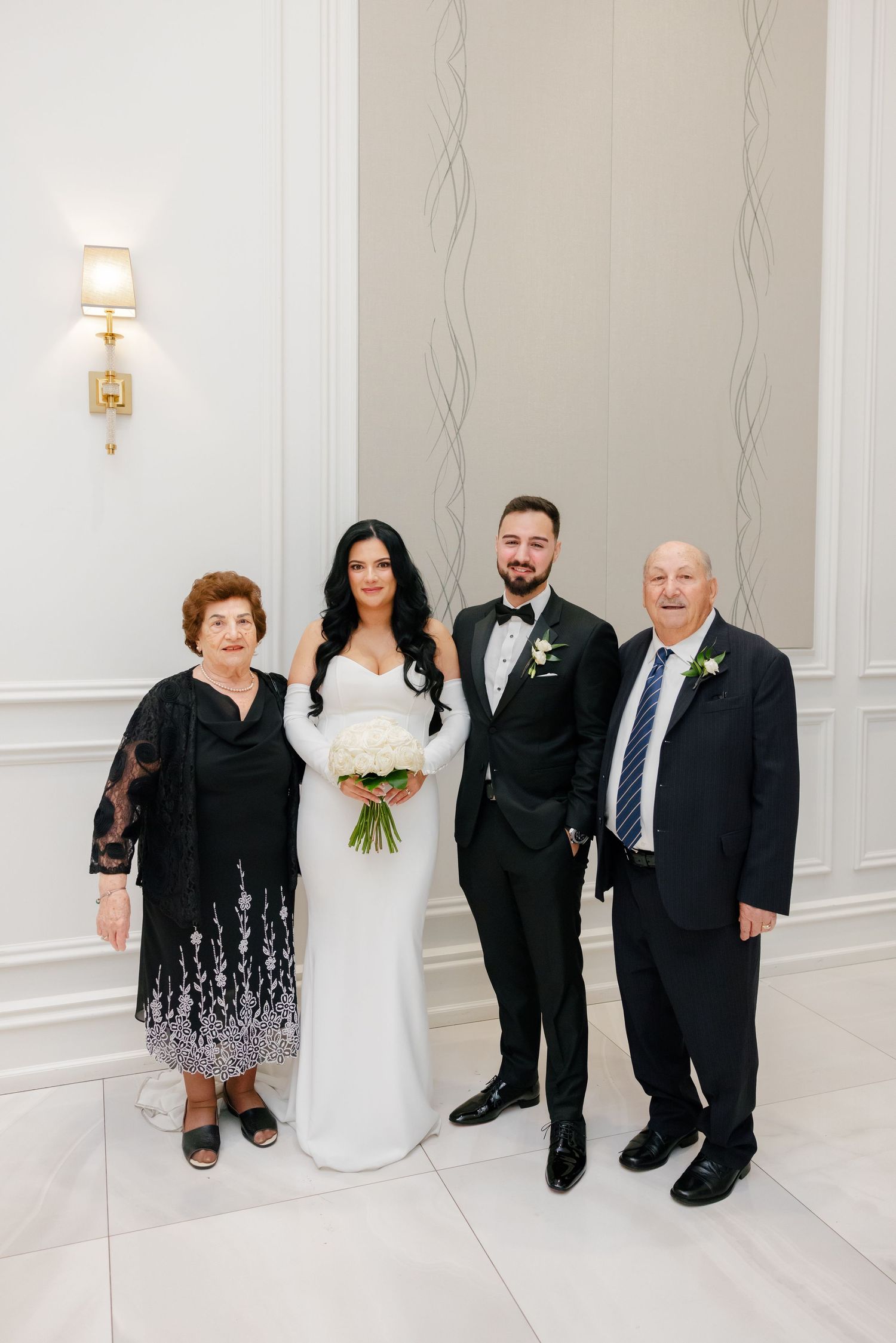 Wedding portrait of newlyweds with older relatives in formal attire against white wall.