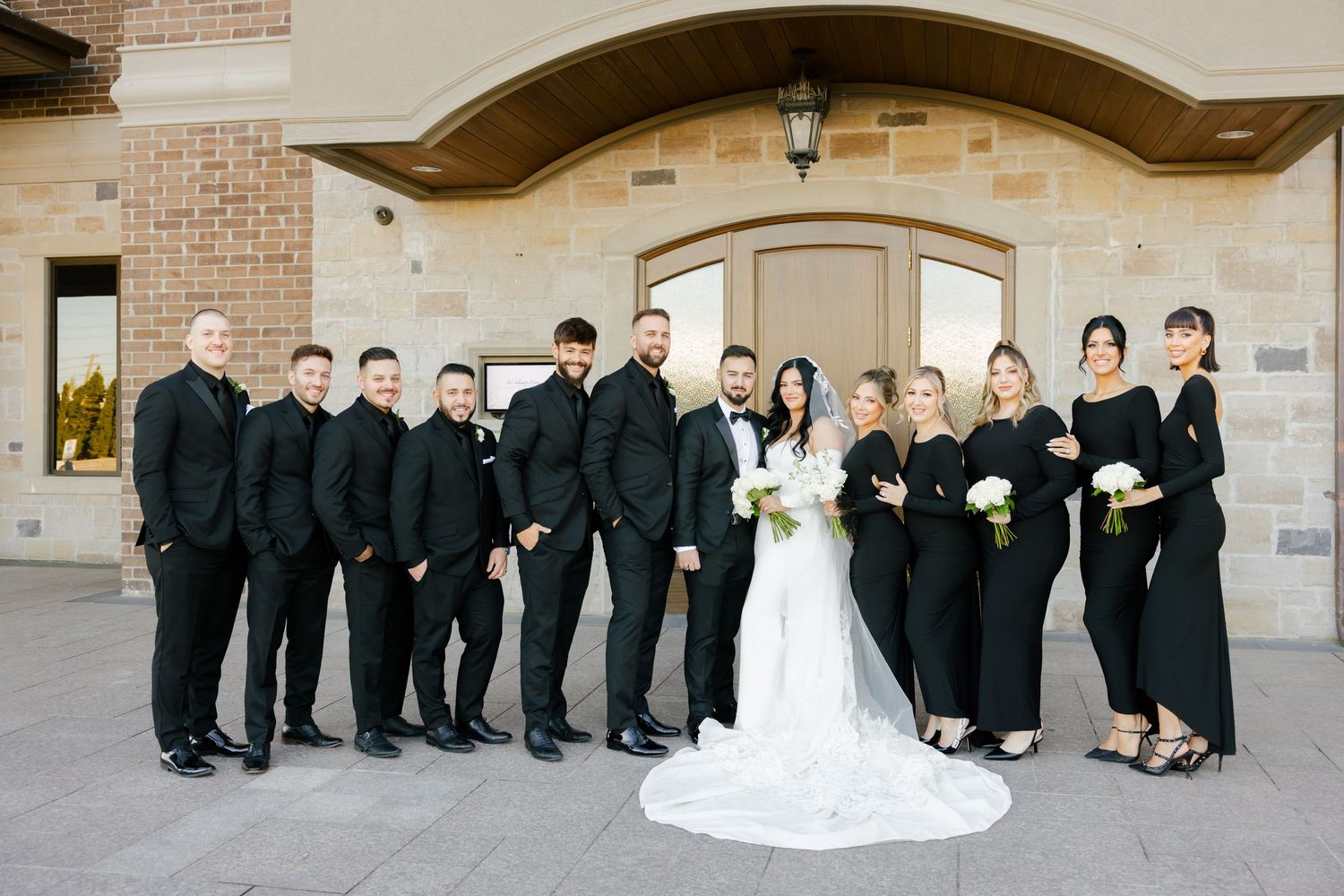 Elegant wedding party wearing black and white formal attire poses in front of a stone archway.