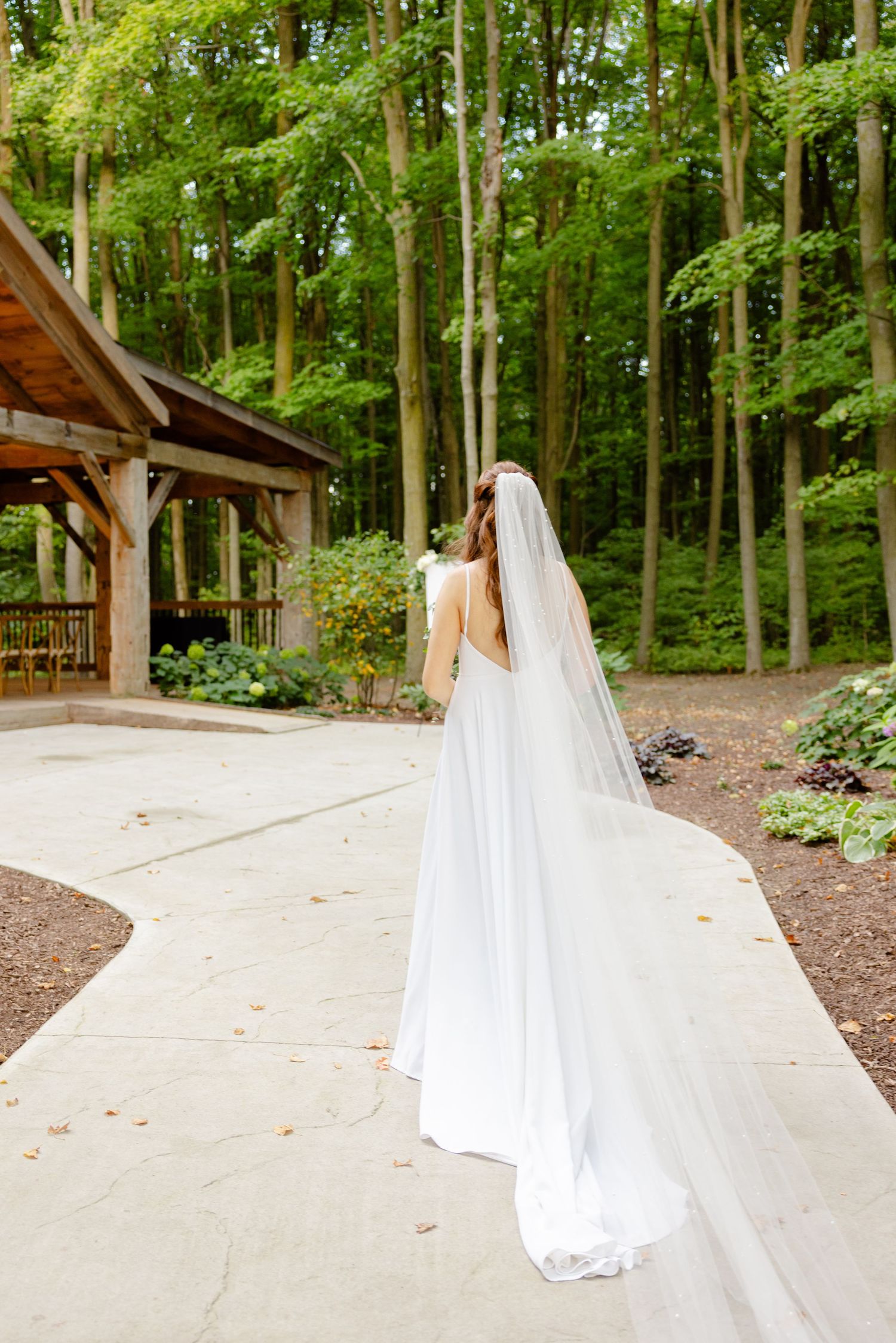 Bride in flowing white dress and veil stands on pathway near rustic wooden pavilion in forest setting.