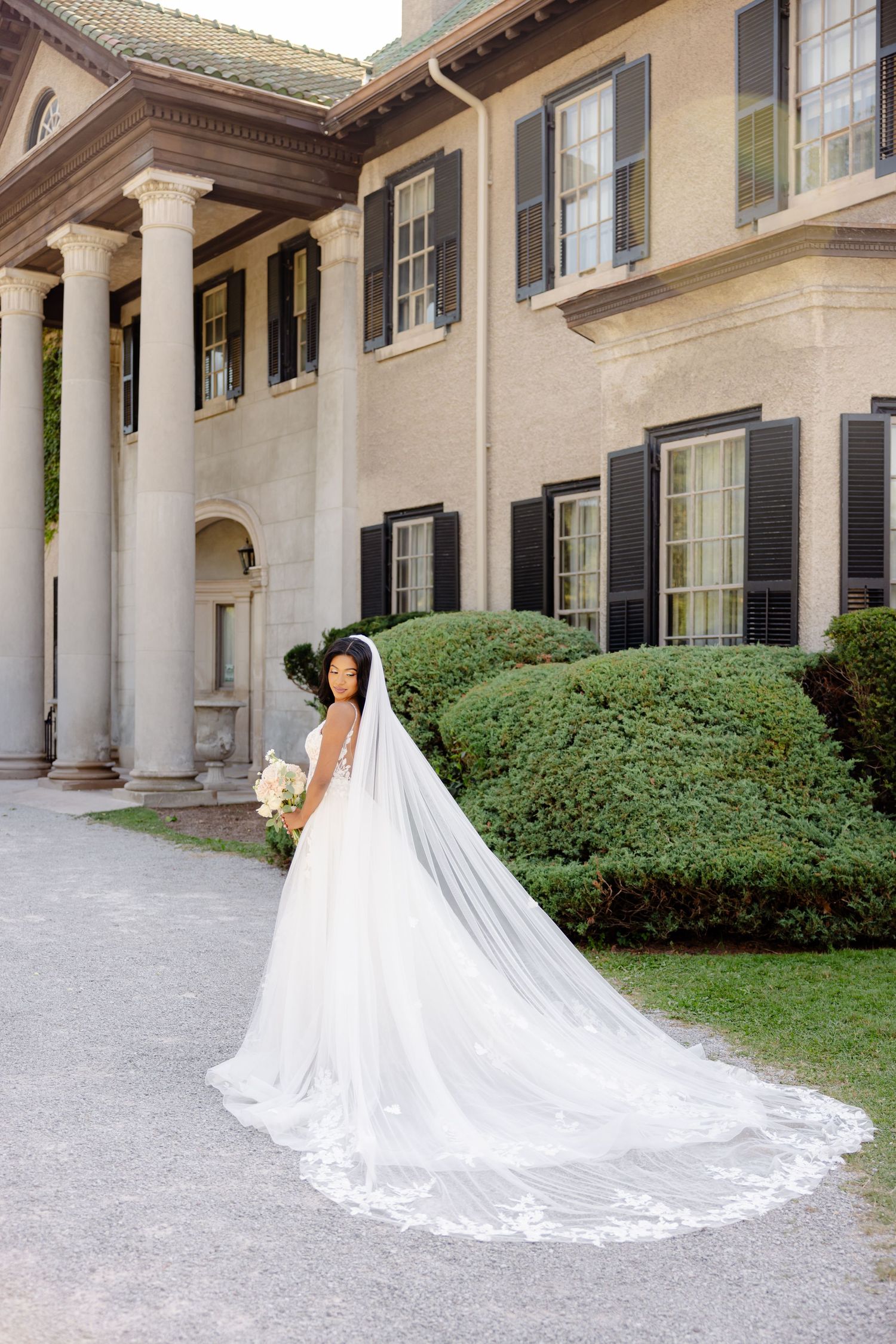 Bride in white gown with dramatic cathedral veil poses outside elegant colonial building.