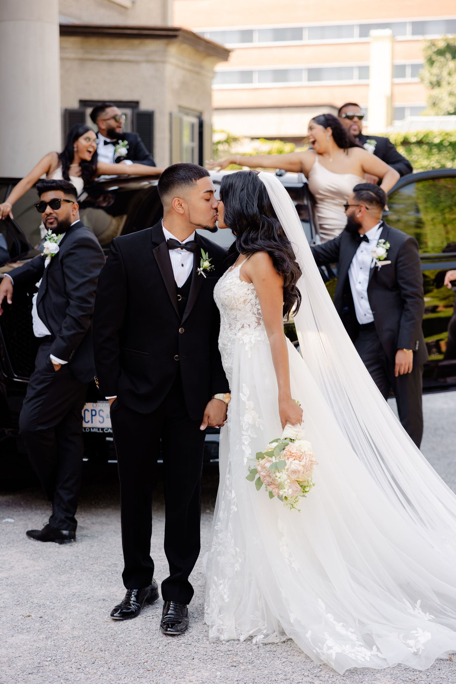 Wedding party in formal attire gathers around a luxury car outside grand building entrance.