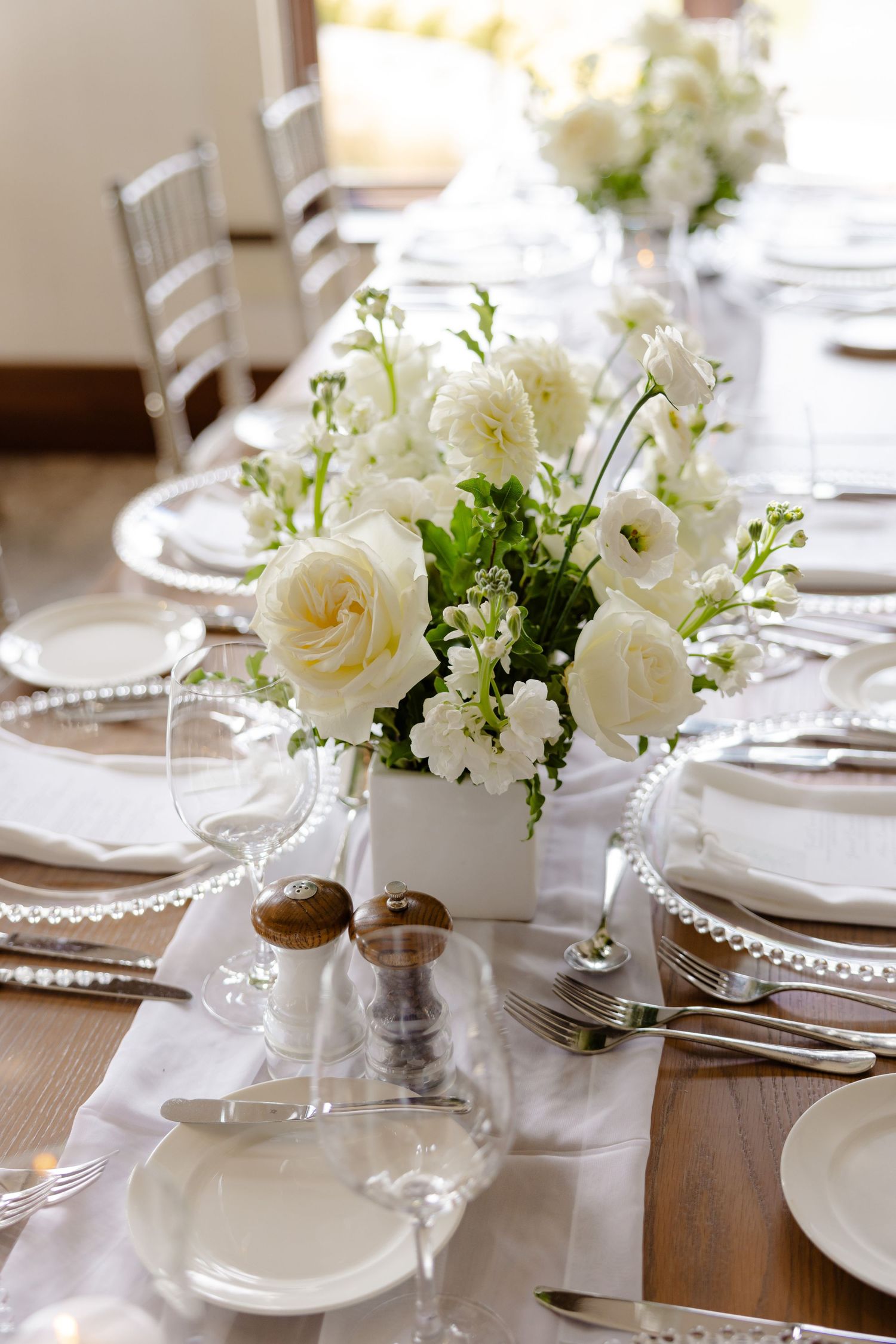 Romantic white rose and greenery centerpiece on a formal dining table with place settings and salt shakers.