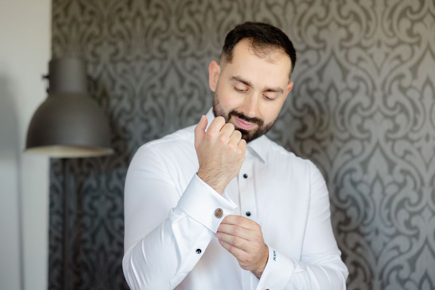 Person in white dress shirt adjusting cufflinks while getting ready against grey patterned wall.