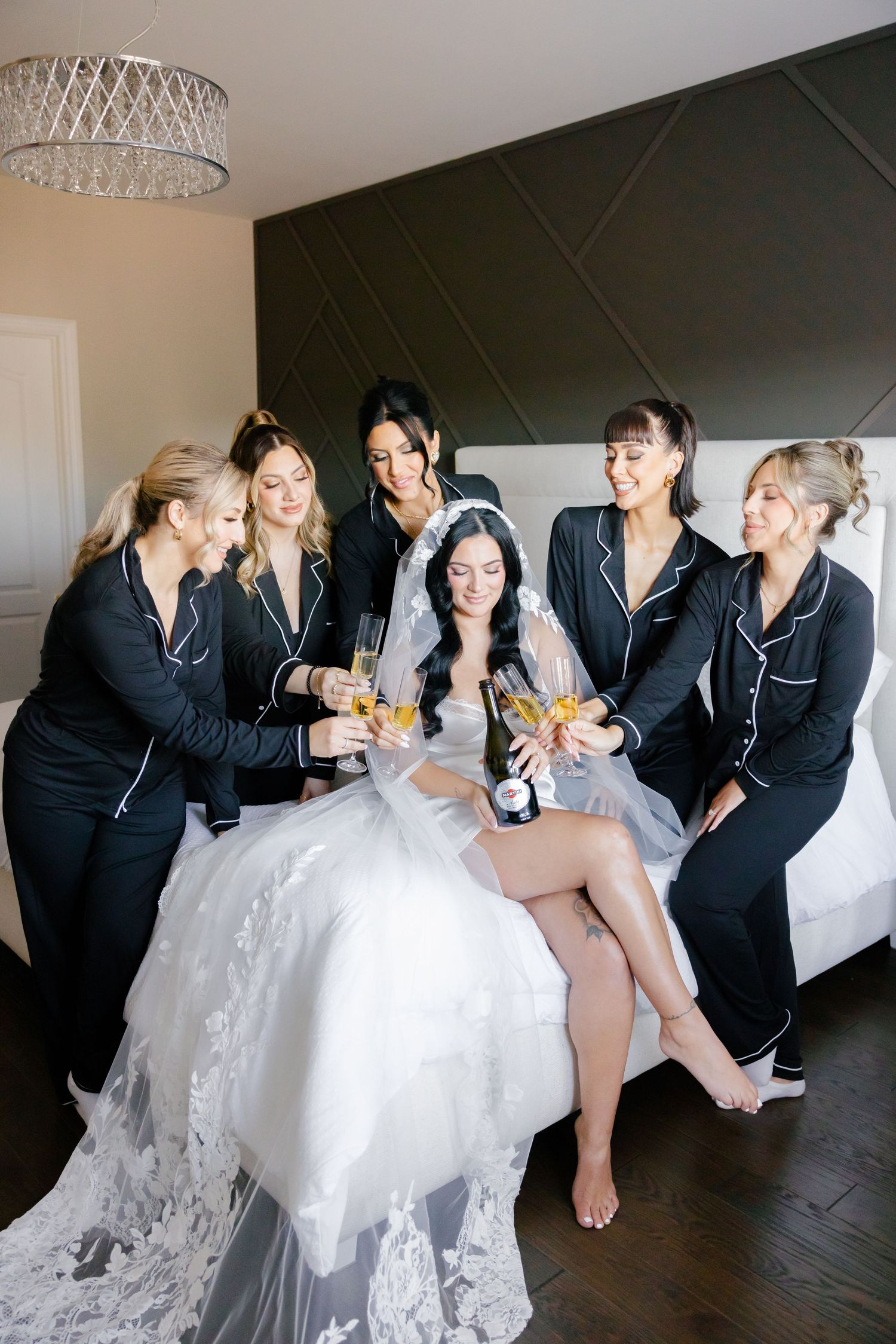 Bridal party toasts with champagne on bed, wearing matching black sleepwear.