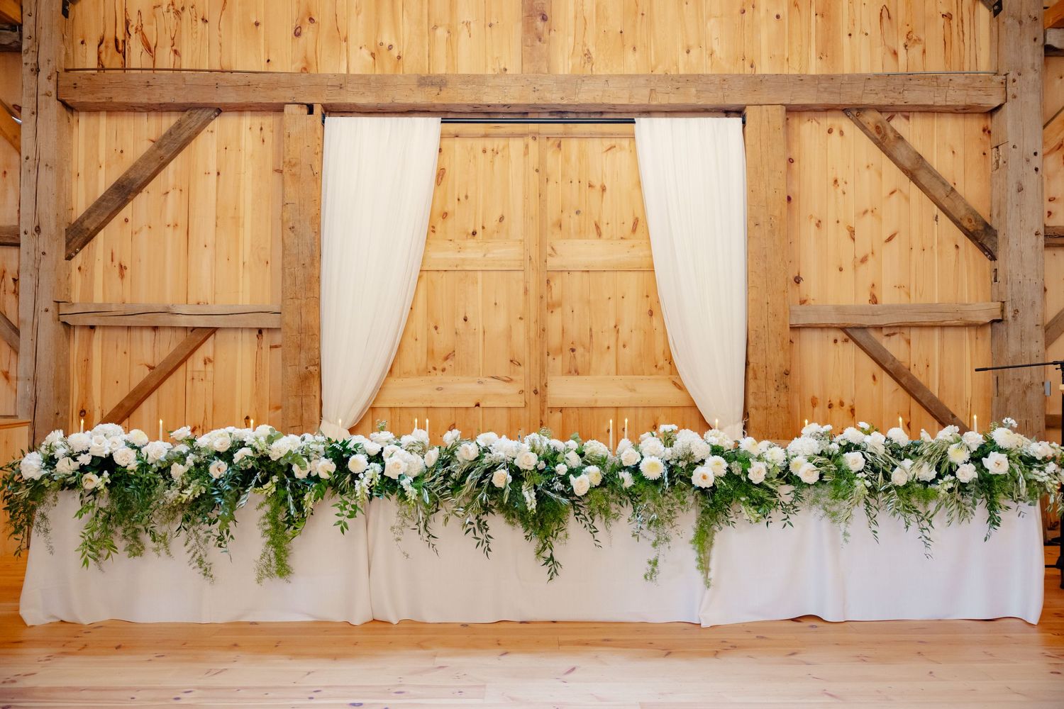 White floral and greenery garland decorating a wooden backdrop with flowing white curtains.