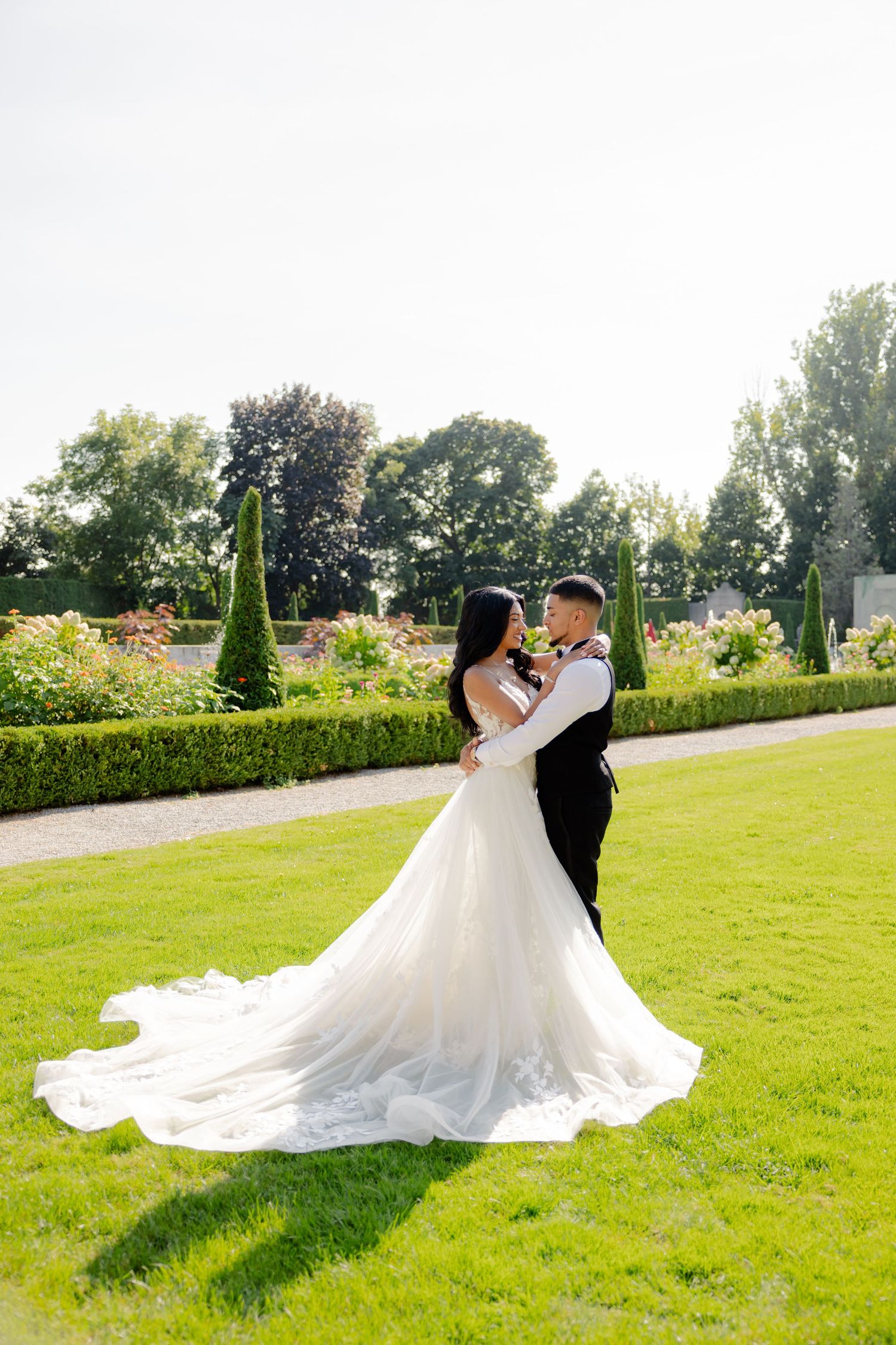 A couple in wedding attire embraces on perfectly manicured lawn with topiary garden backdrop.