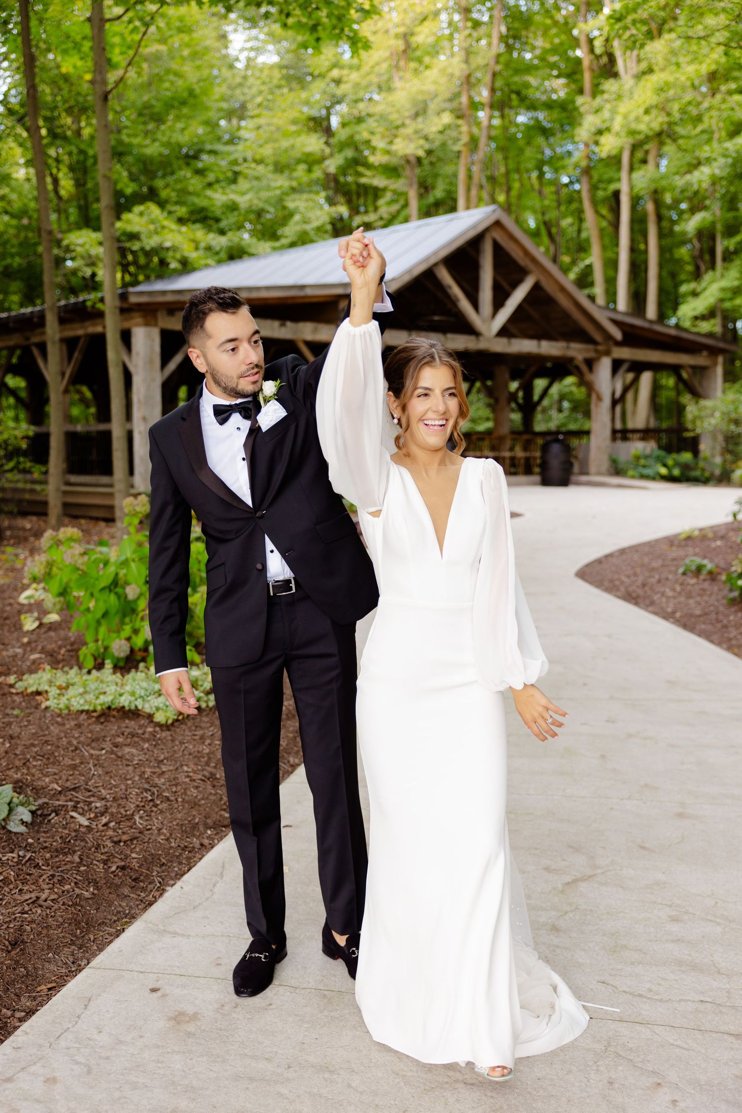 Joyful couple celebrates with raised hands on a forest path by a wooden pavilion.