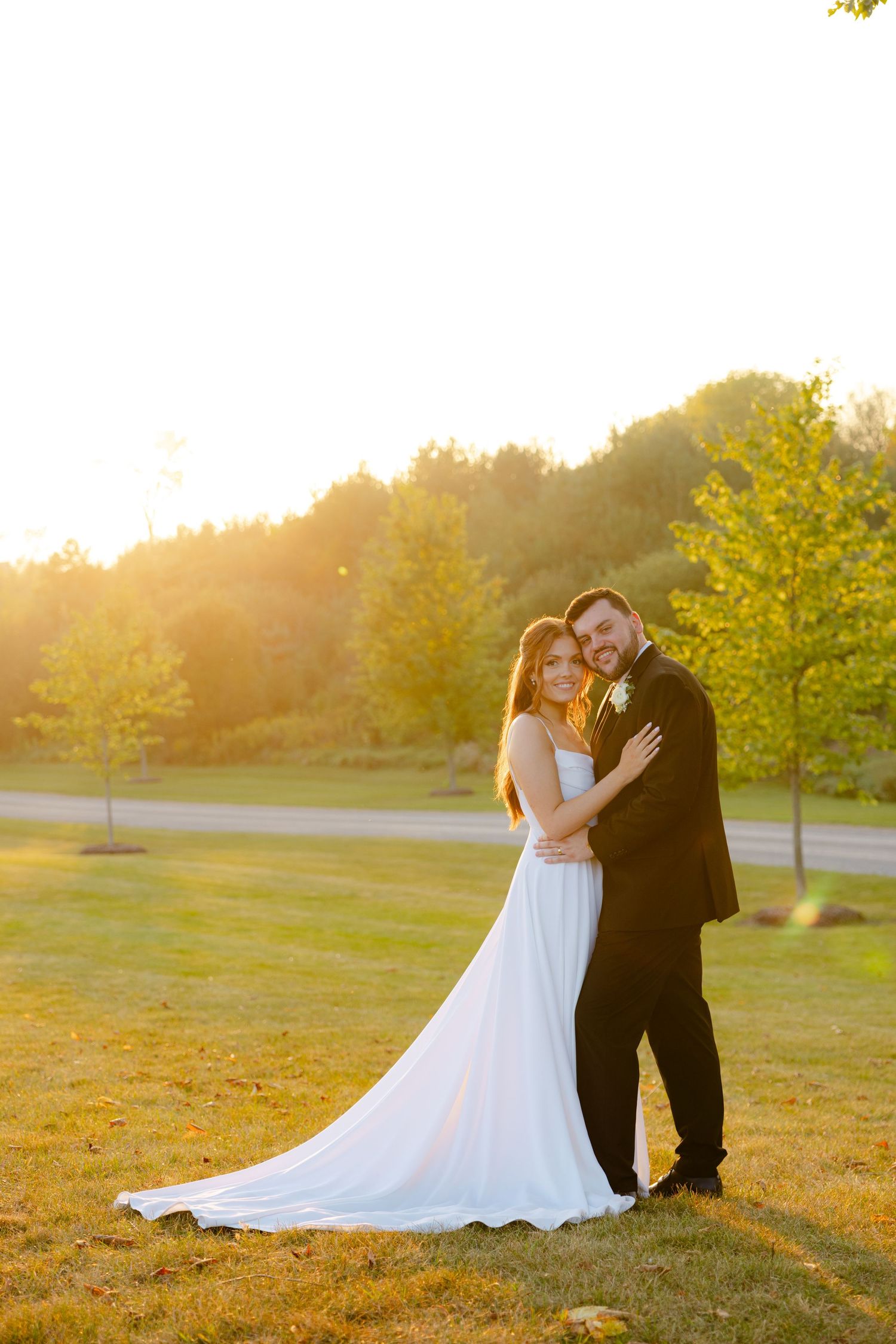 A bride in white gown and groom in black suit pose together on a hillside during sunset.