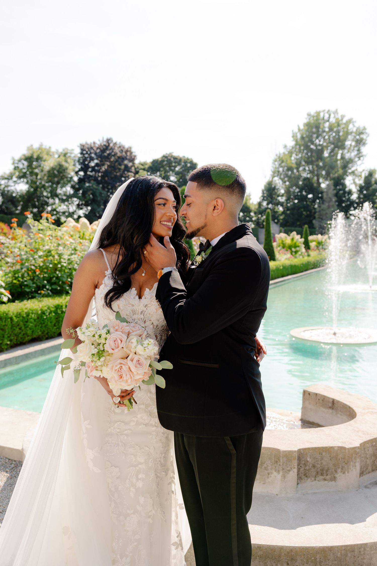 A couple shares an intimate moment in a garden setting with a fountain backdrop.