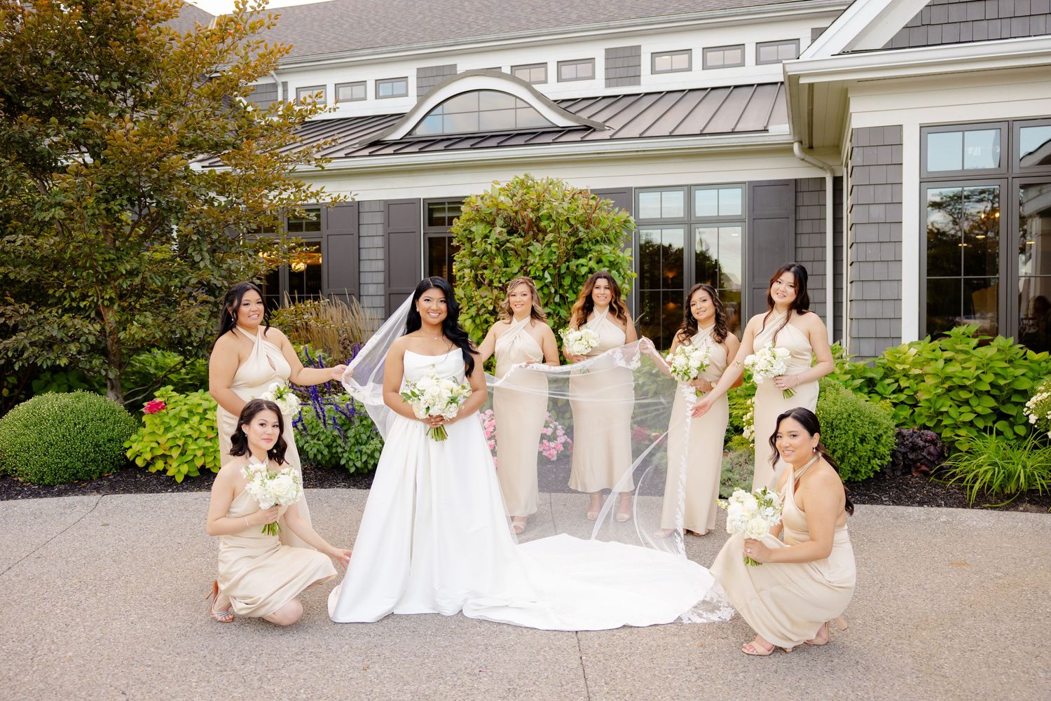A wedding party poses outside a gray building with bridesmaids in champagne dresses and a long white gown.
