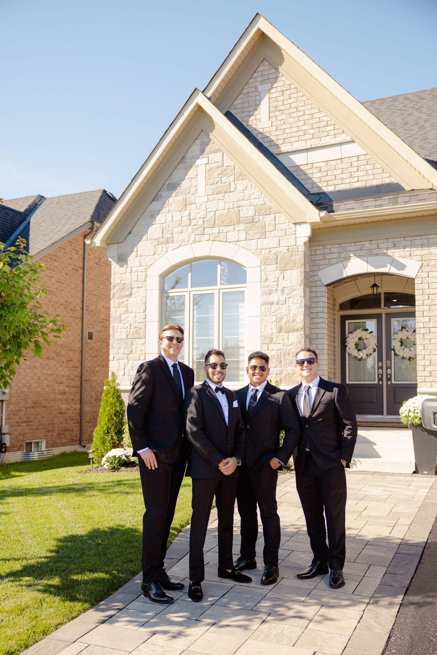 Groomsmen in black suits and sunglasses pose in front of a limestone house on a sunny day.