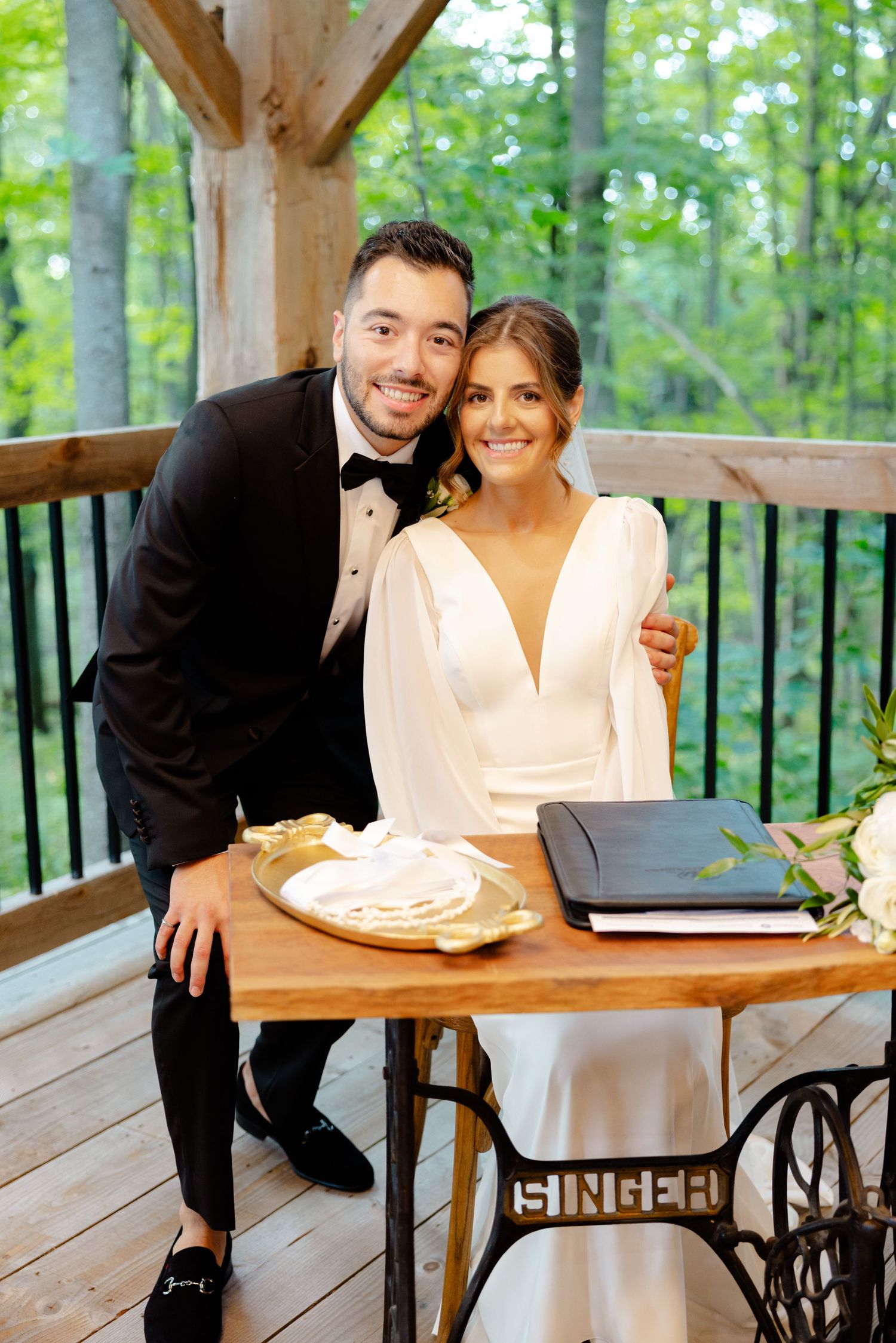 Newlyweds pose by antique Singer sewing table in rustic forest pavilion setting.