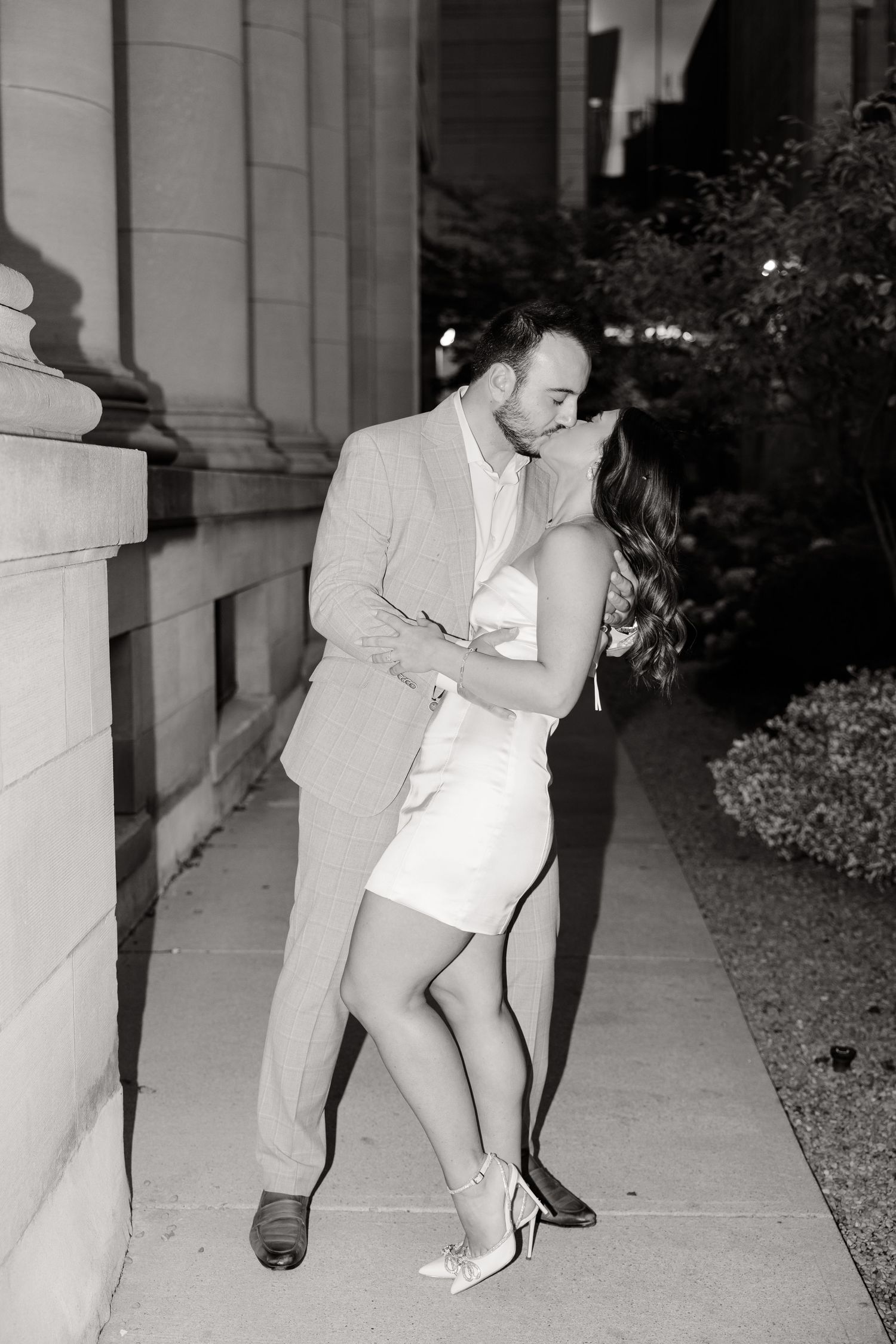 A romantic black and white photo of a couple embracing near stone columns.