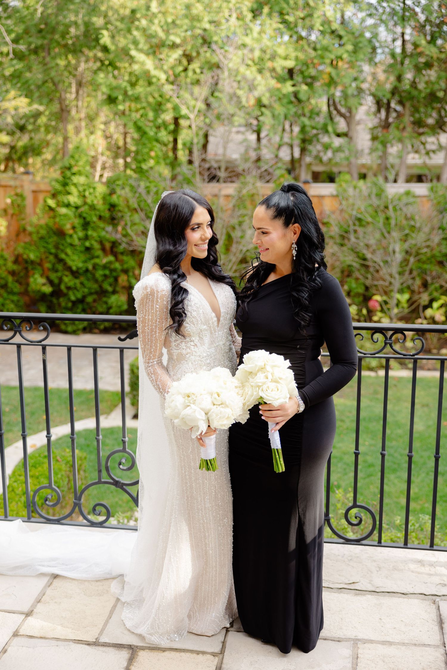 Two women in formal attire share a moment on stone patio, one in white bridal gown, other in black dress.