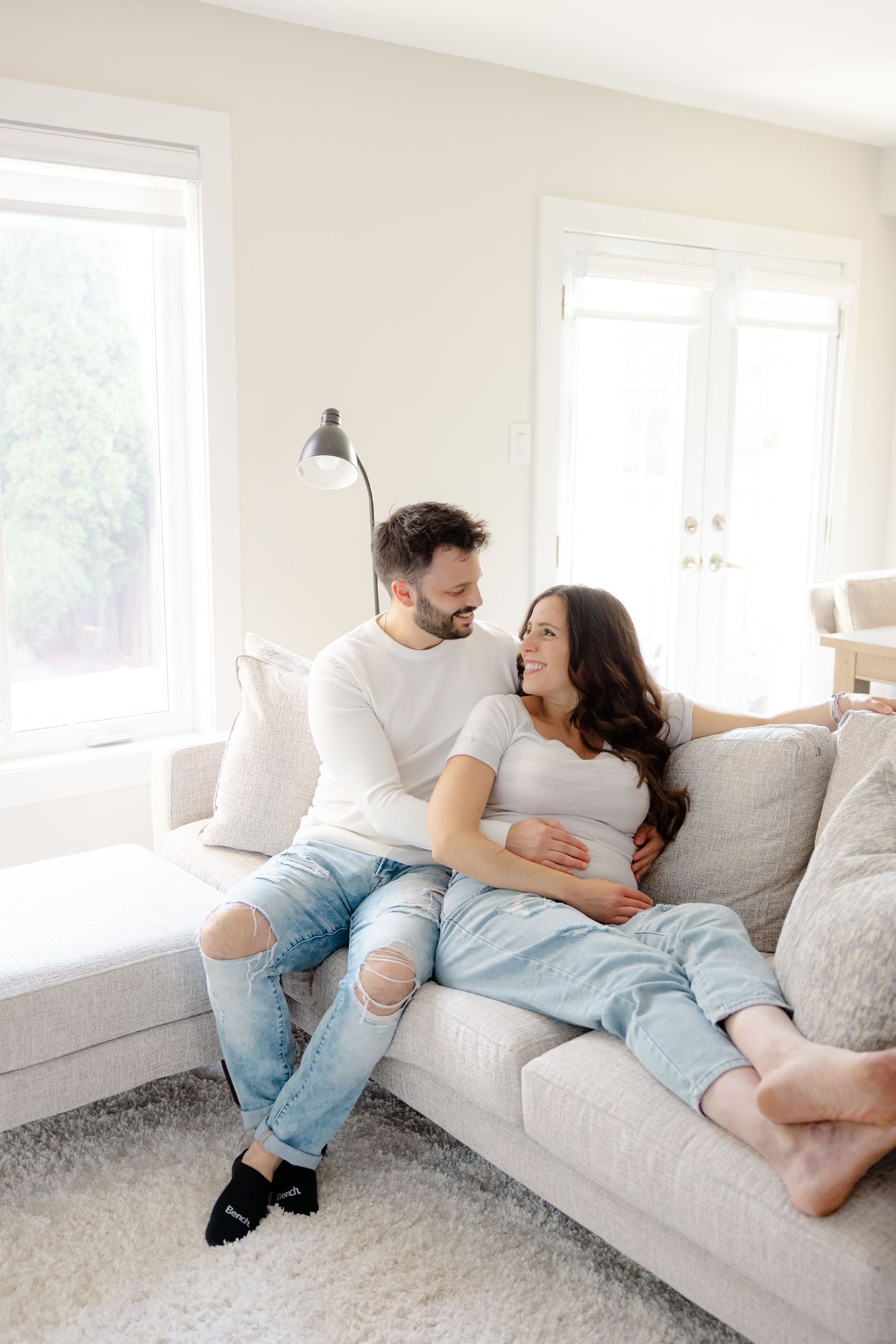 Couple relaxing on a white couch in a bright, airy room with large windows and sunlight streaming in.