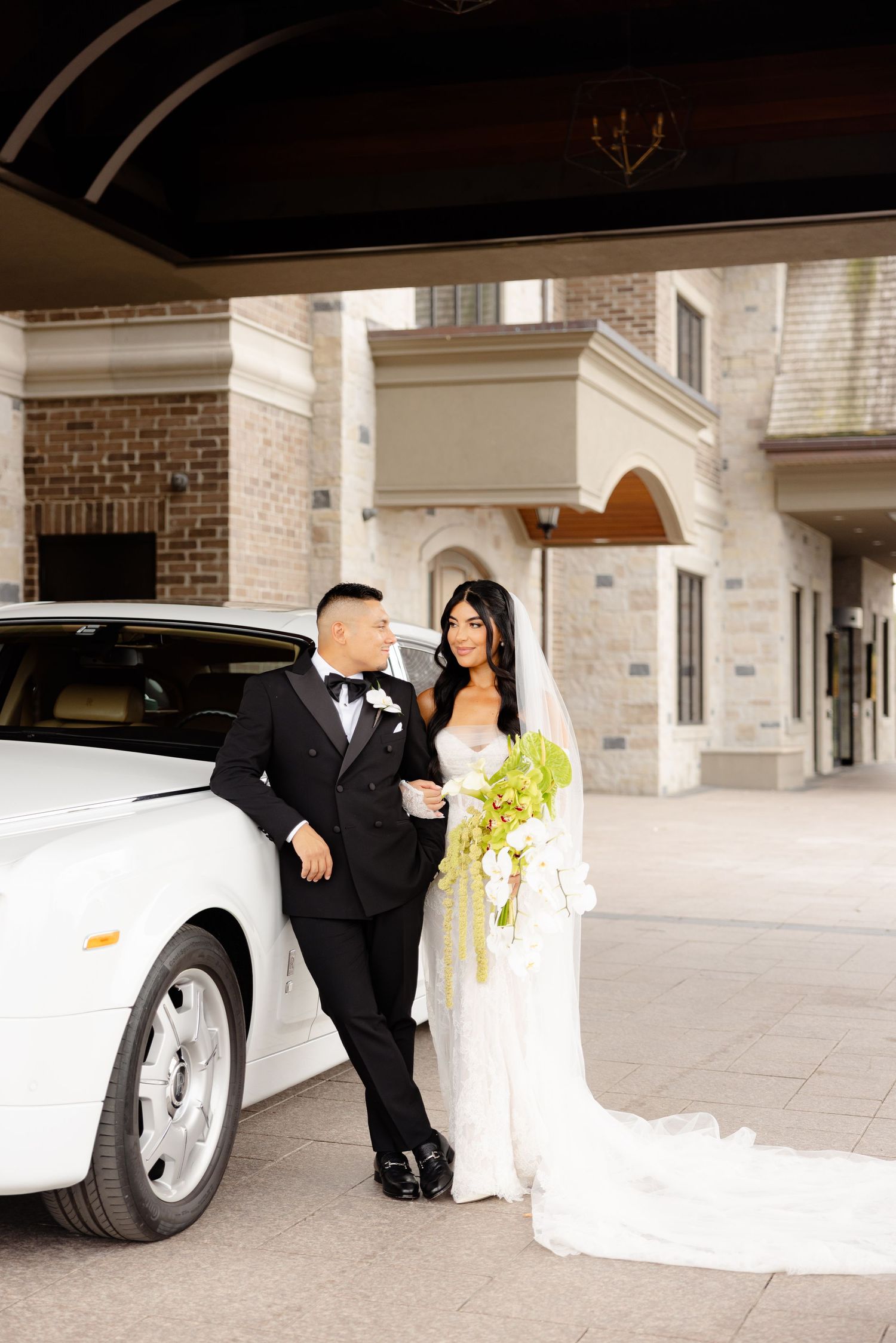 A couple in wedding attire pose elegantly next to a luxury white limousine in front of a brick building.