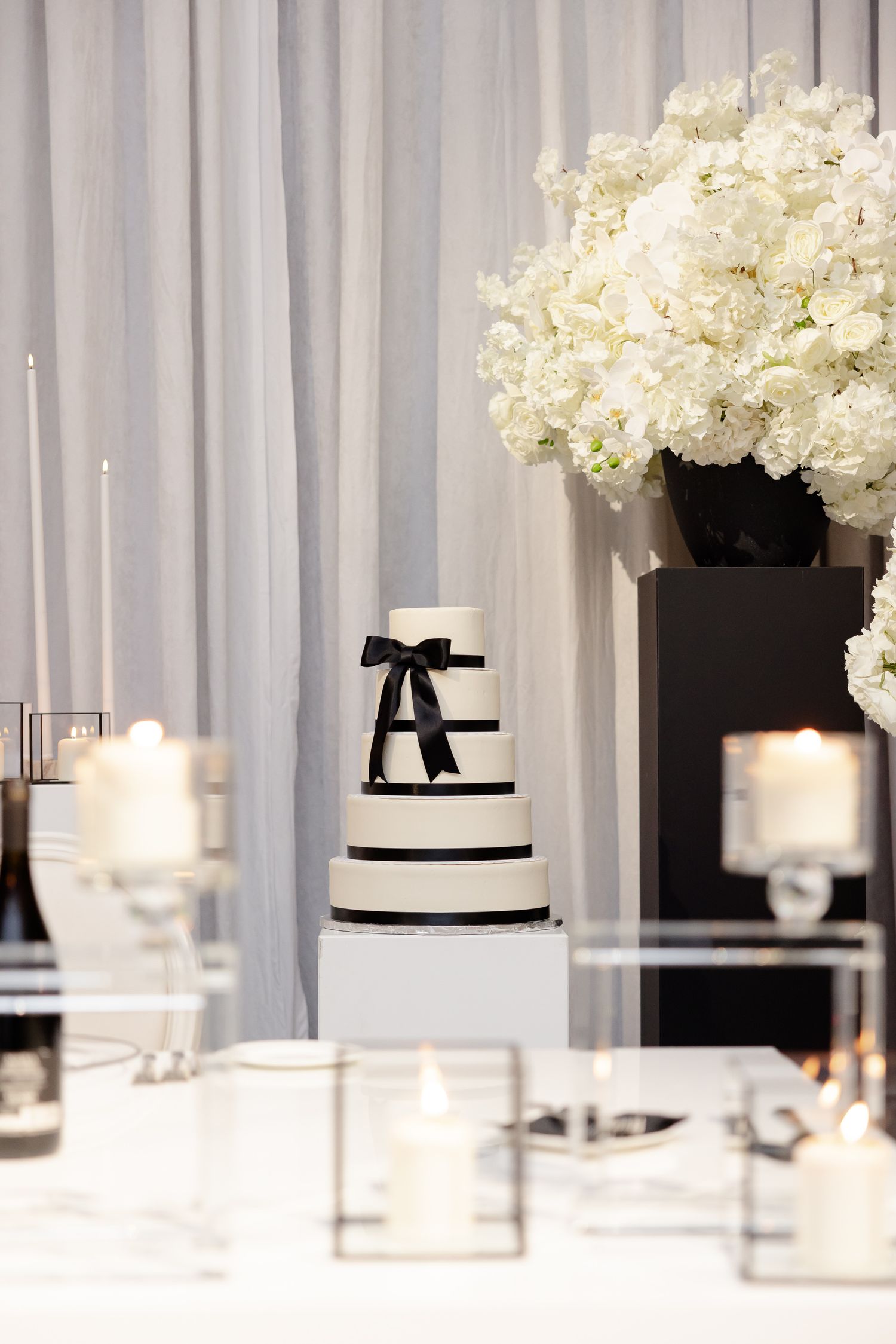 An elegant black and white tiered wedding cake with a bow on top, surrounded by white hydrangeas and glowing candles.