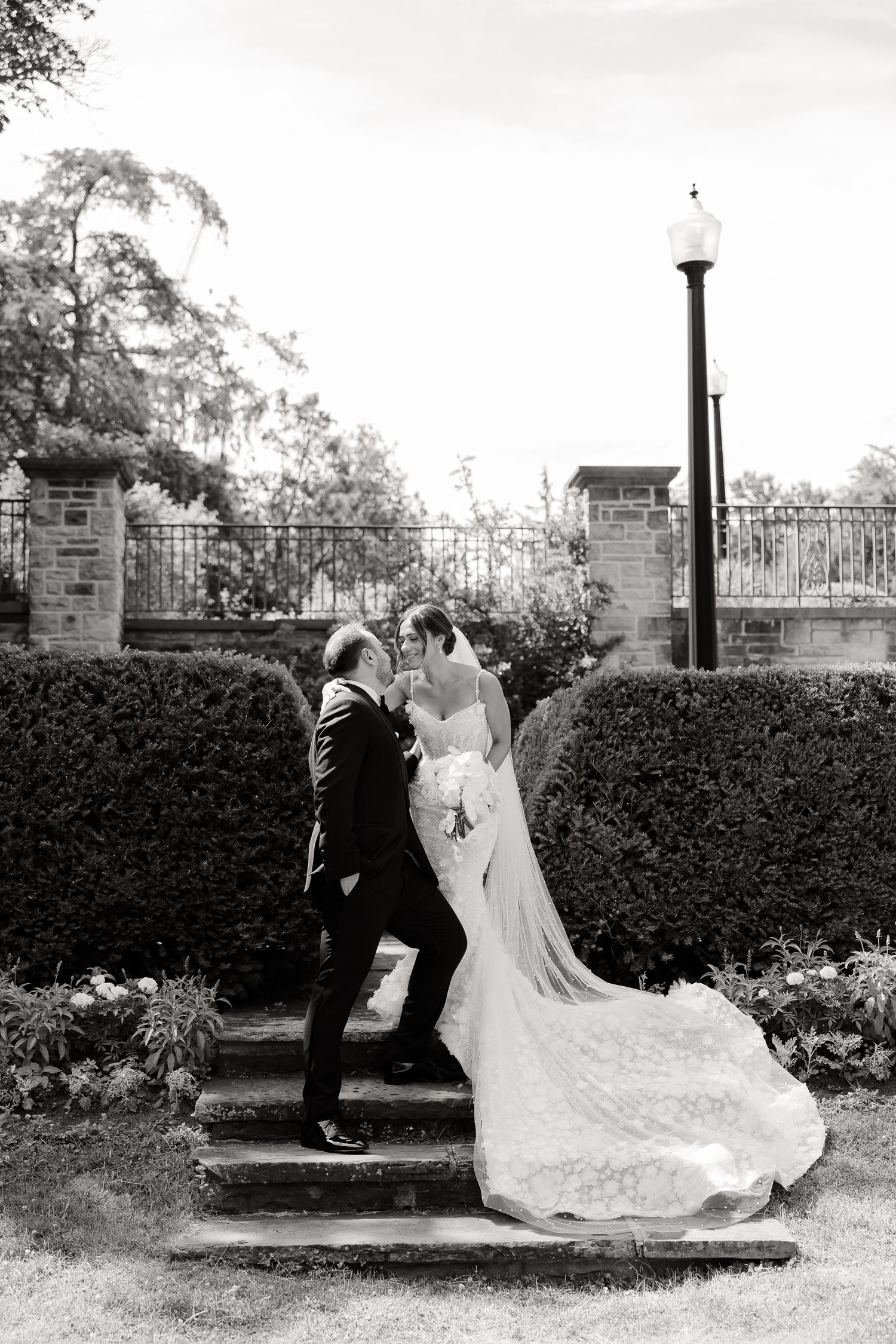 A black and white romantic wedding photo captured on garden steps with decorative lampposts and hedges in the background.