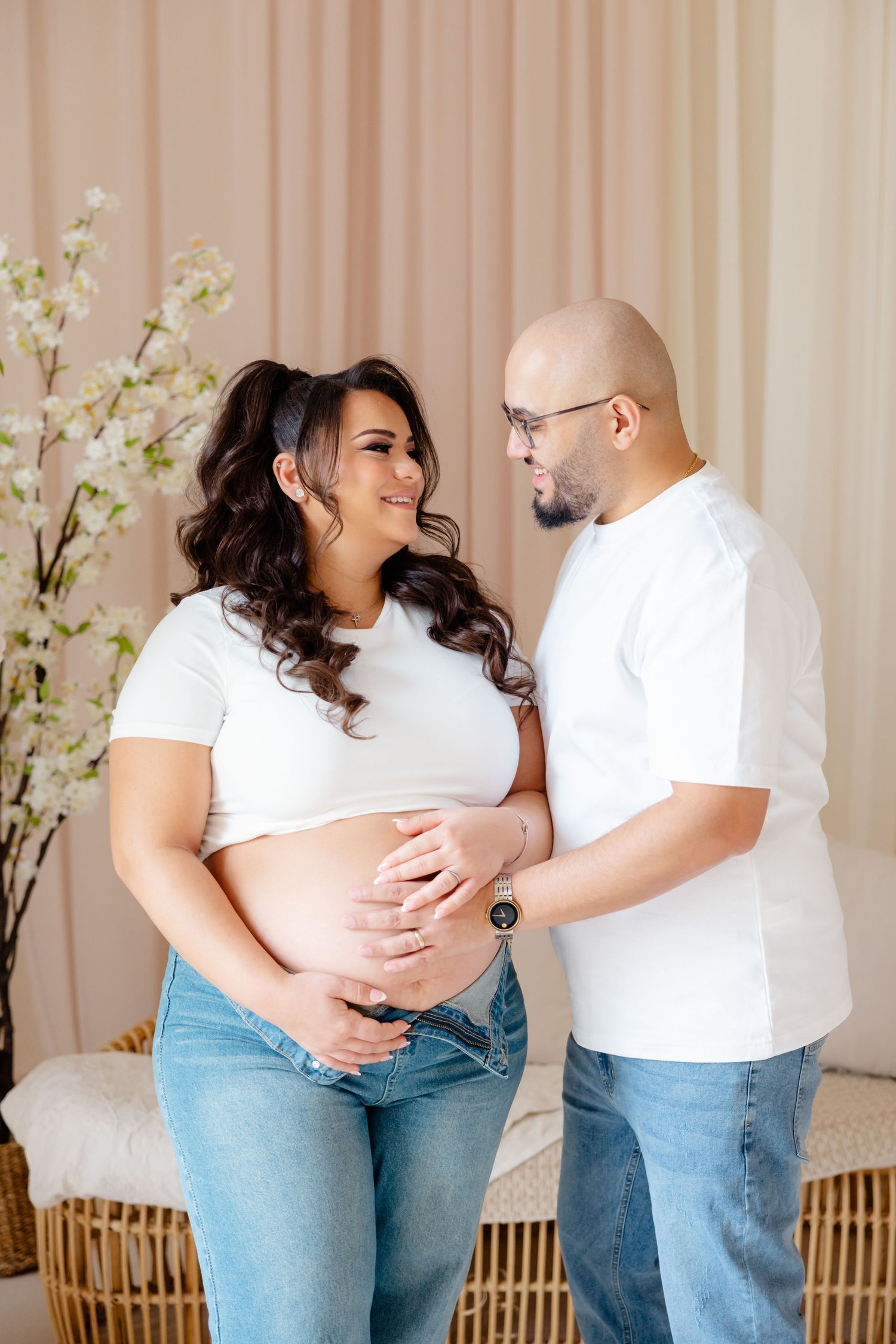 Expecting couple in matching white shirts and jeans share an intimate moment in front of floral decor at home.