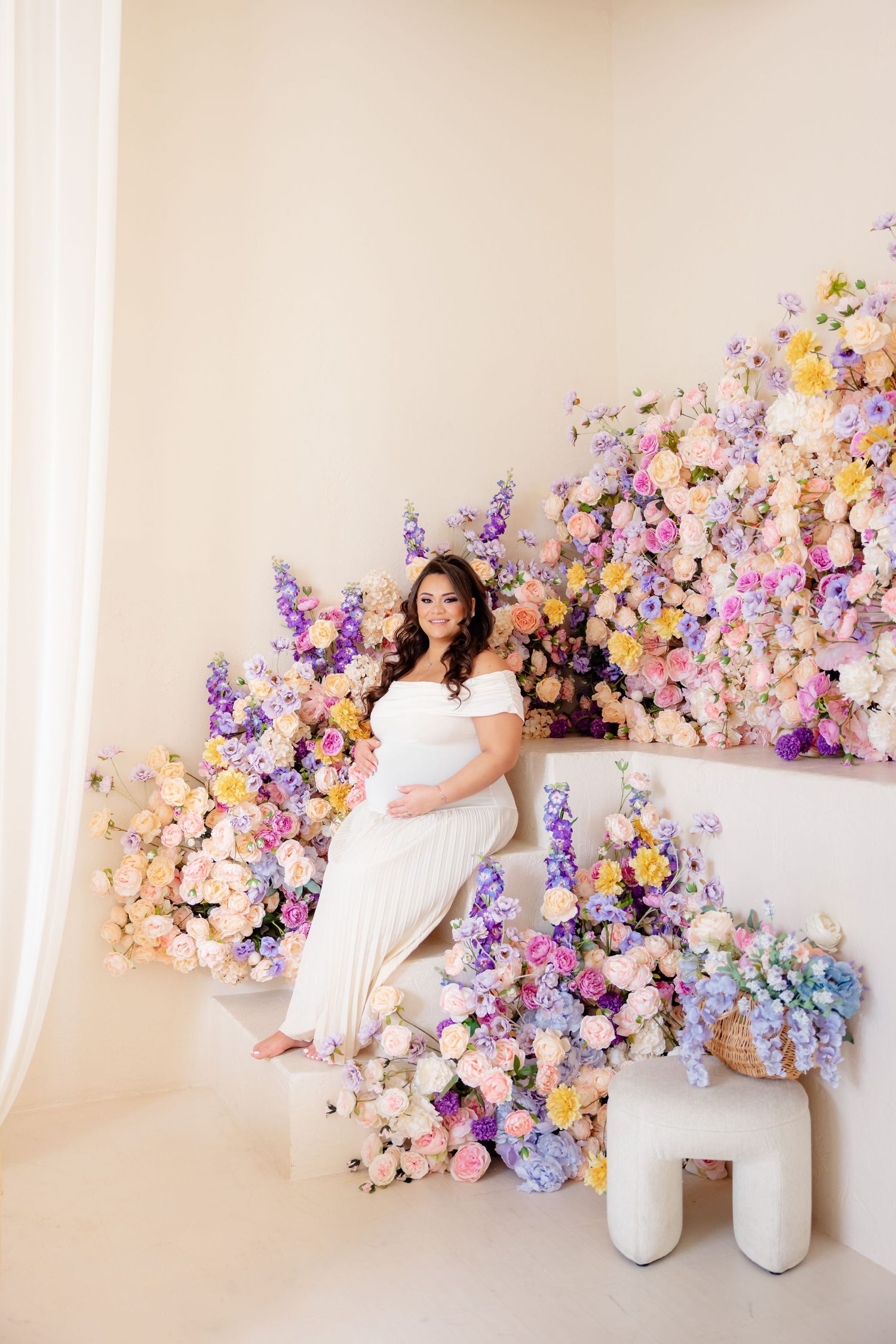 Dreamy maternity photo setup with pastel floral arrangements of pink, purple and yellow blooms against cream walls.