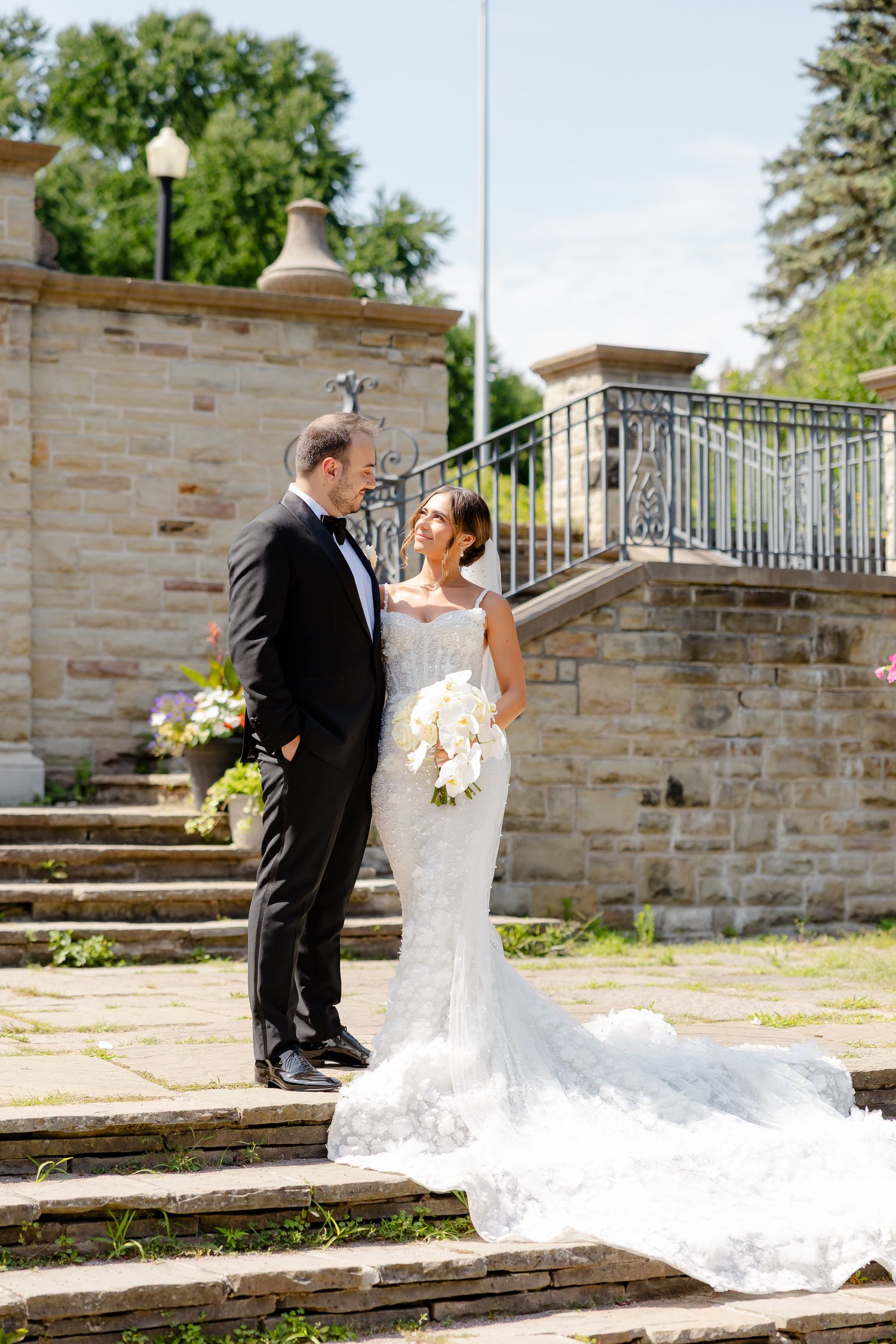 A wedding couple dances together on stone steps outdoors on a sunny day near a stone wall.