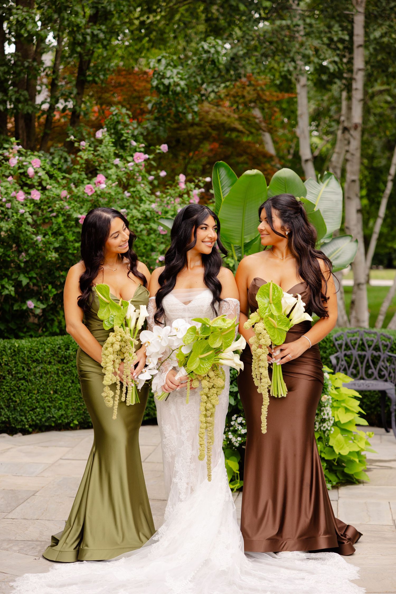 Wedding party poses outdoors wearing elegant silk gowns in olive and brown tones with green floral bouquets.