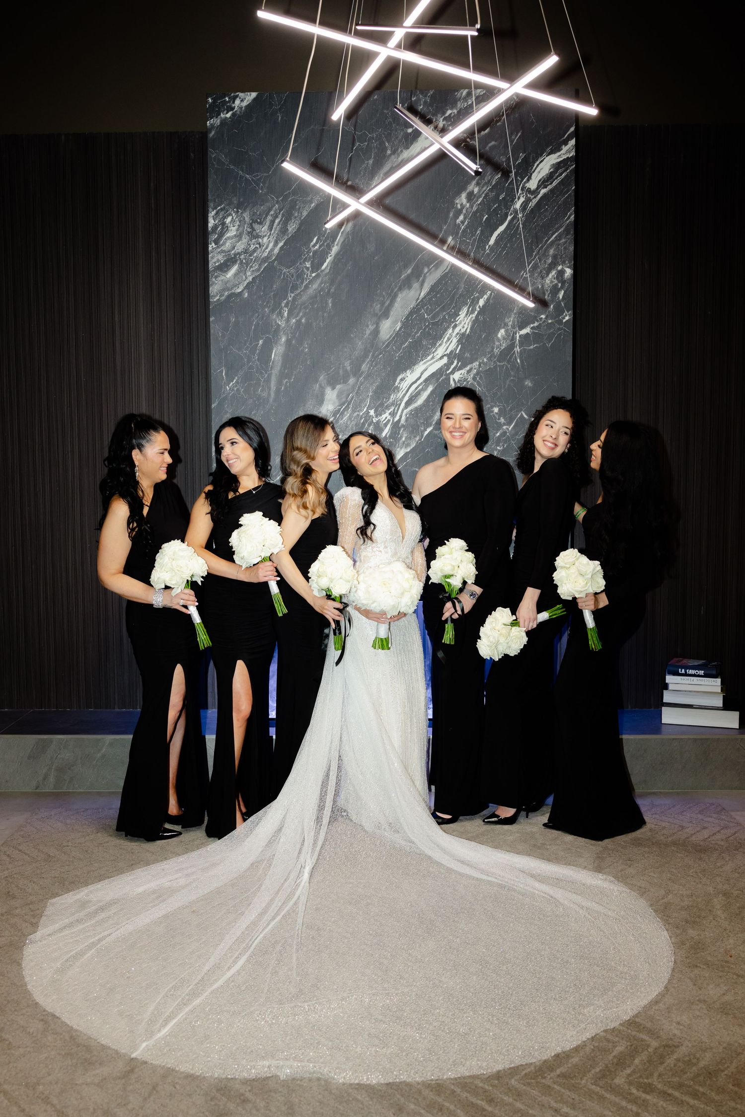 Wedding party in elegant black attire poses with bride under modern lighting fixture.