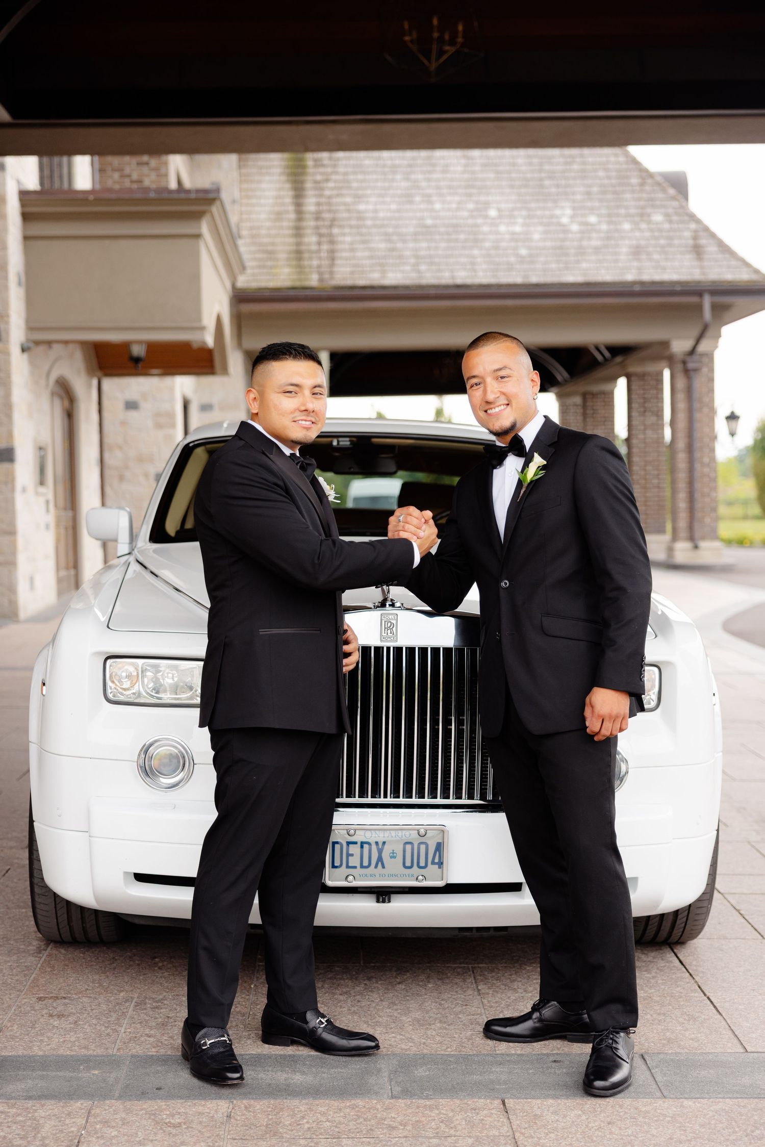 Two people in suits shake hands in front of a white Rolls Royce ghost luxury car at a photoshoot.