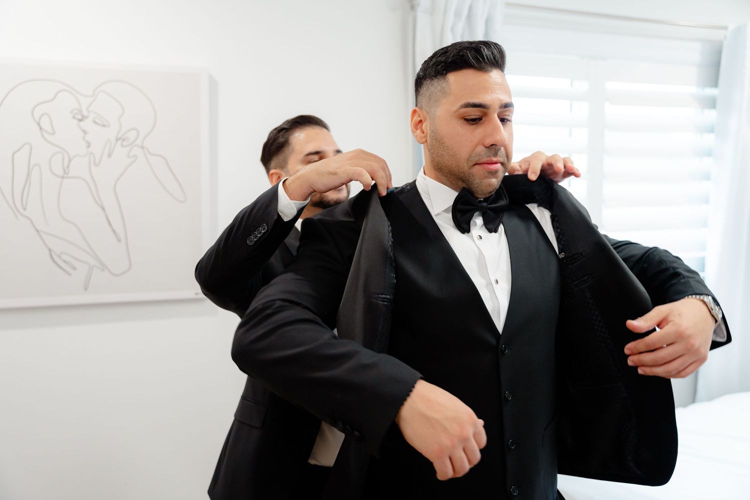 Two people in black tuxedos making final adjustments to their formal attire.