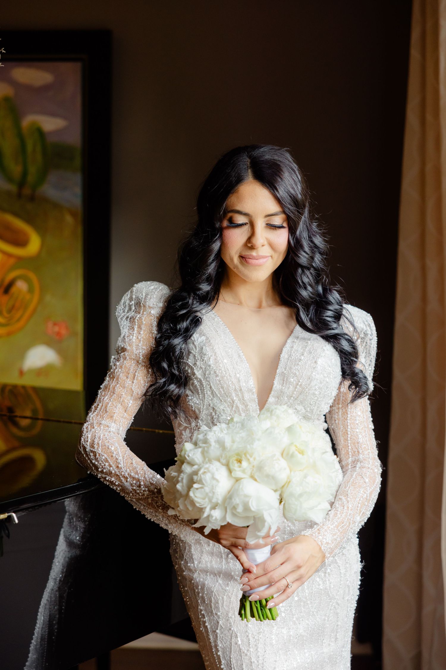Long-sleeved lace wedding gown with plunging neckline and white floral bouquet in elegant interior.