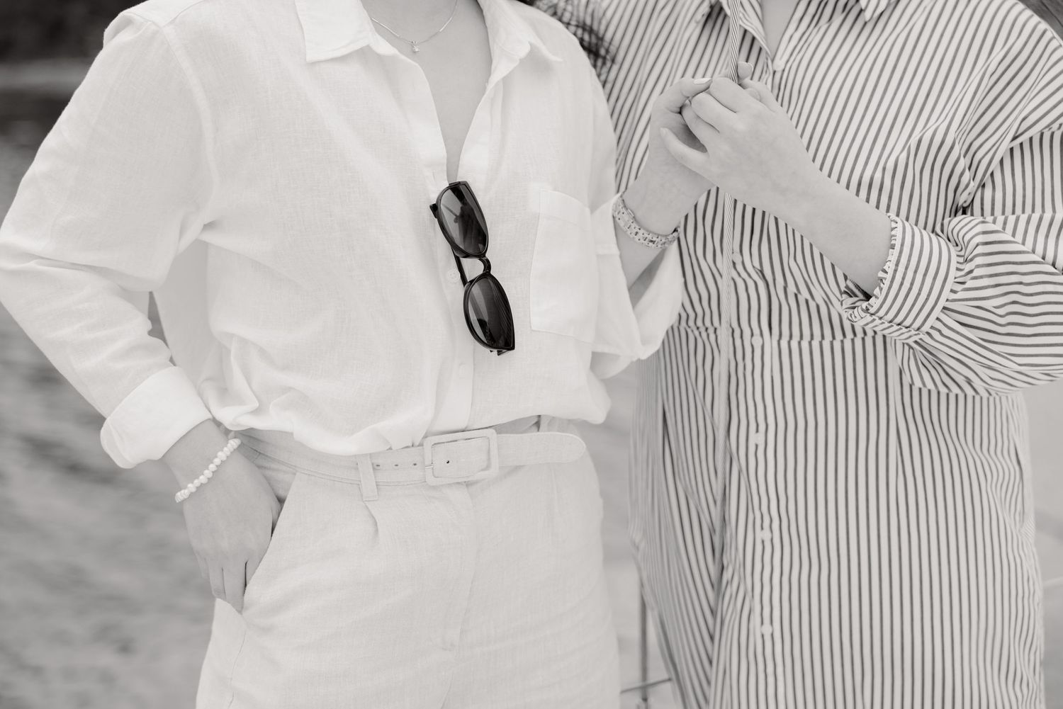 Two people stand together wearing striped and solid white button-down shirts with sunglasses hanging from the collar.