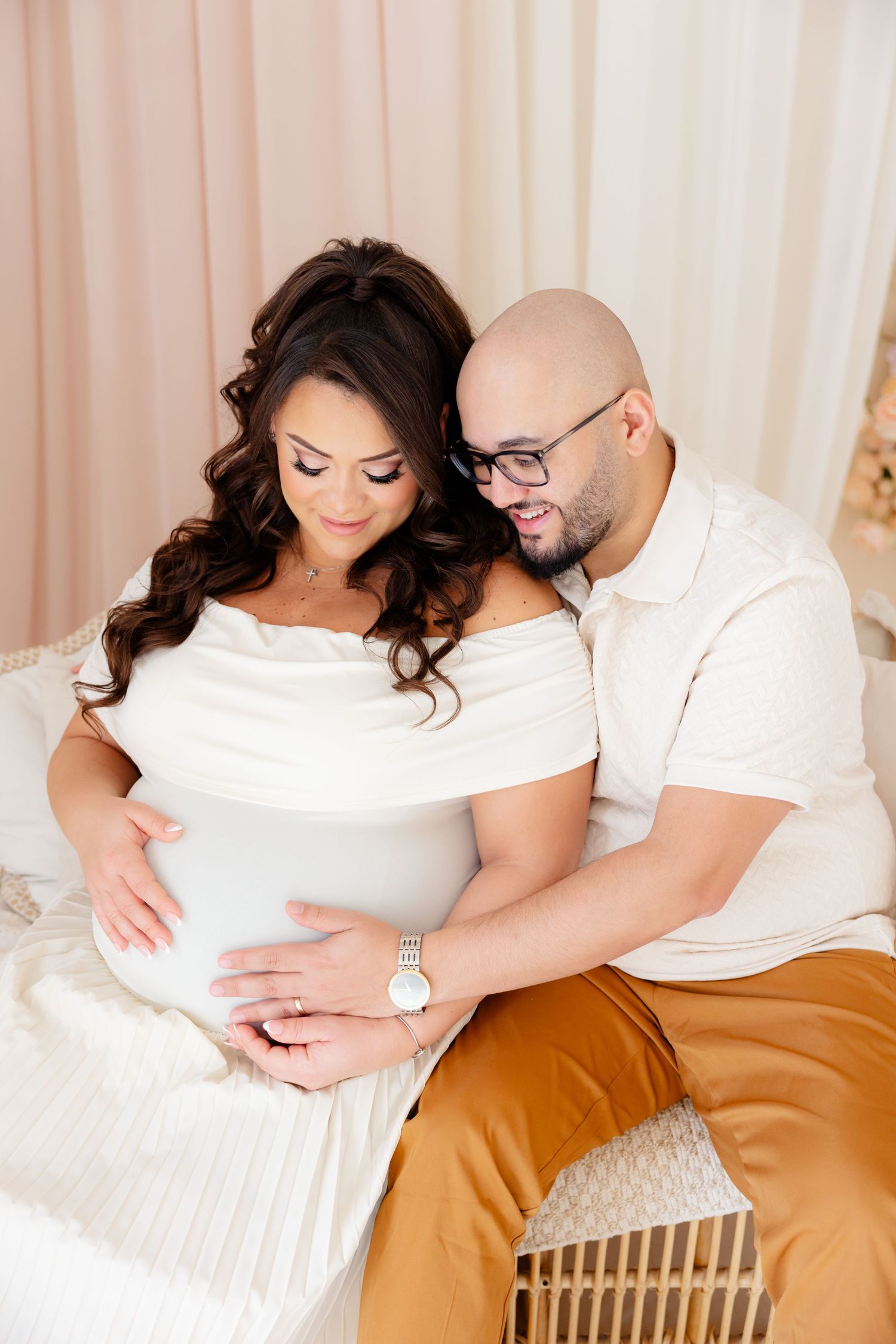 An expecting couple in white outfits share an intimate moment on a sofa while cradling a baby bump.