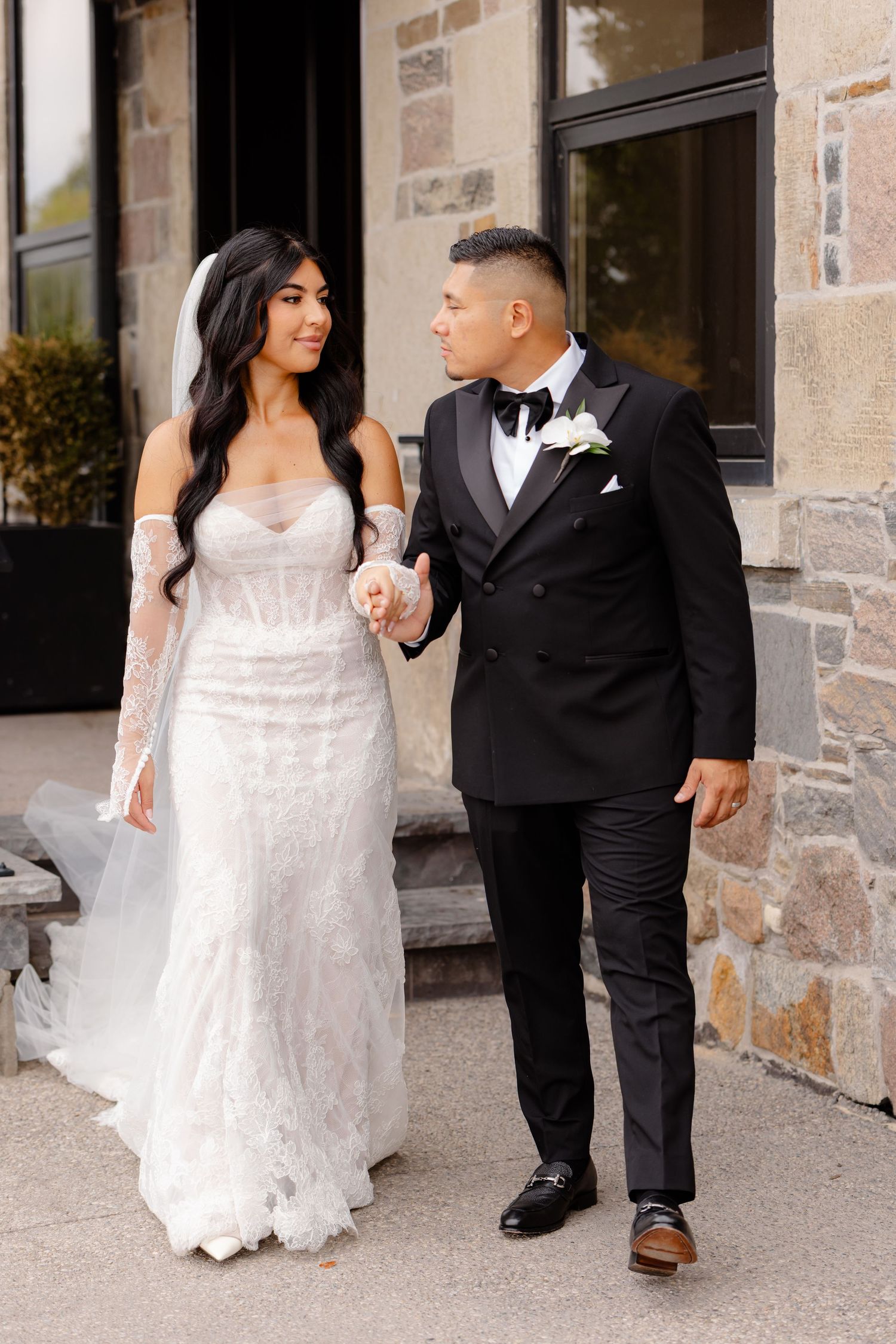 A bride in a white lace gown and groom in a black tuxedo walk together outside a stone building.