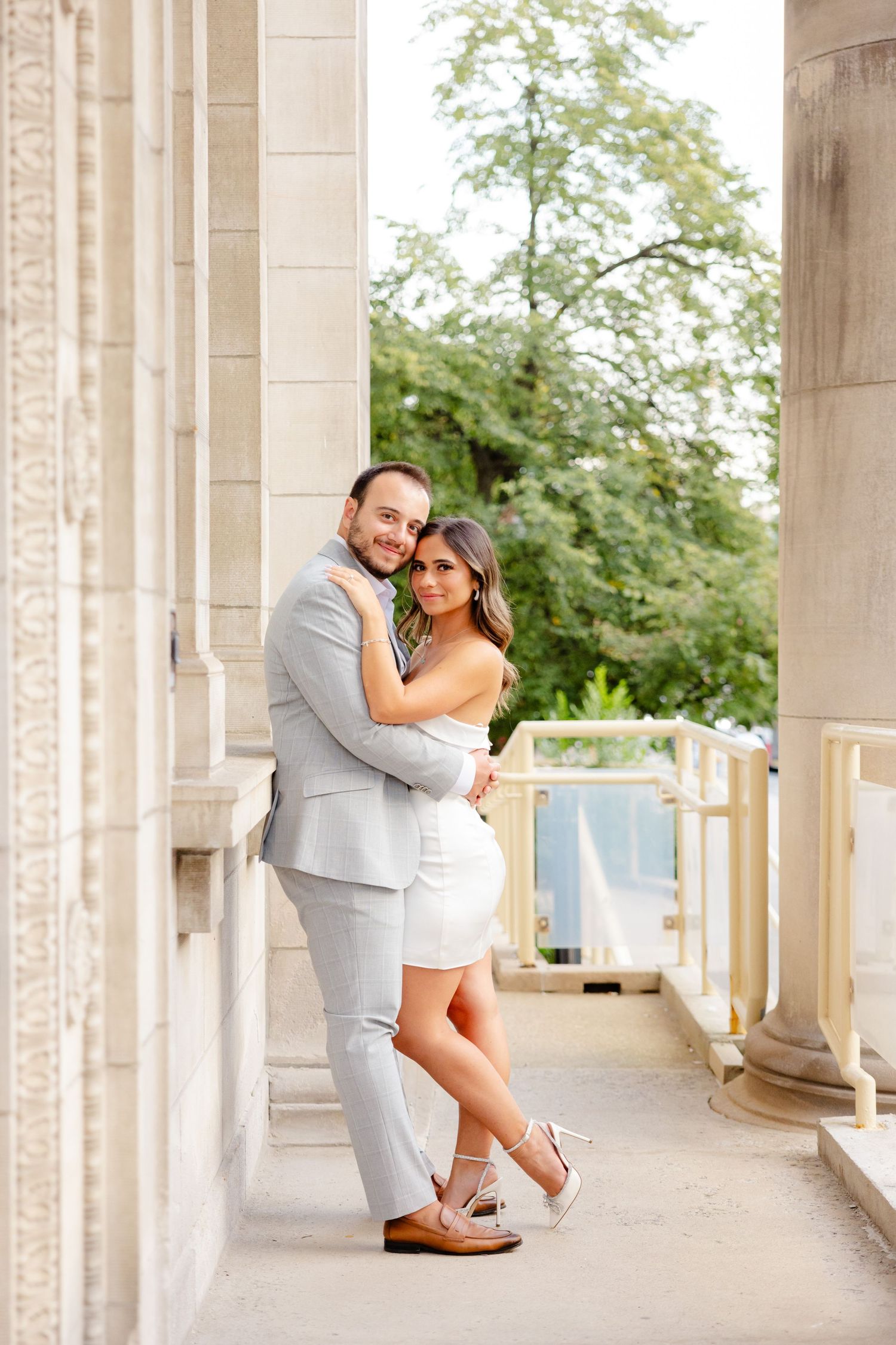 A couple embraces on an elegant covered walkway with white columns and green foliage.