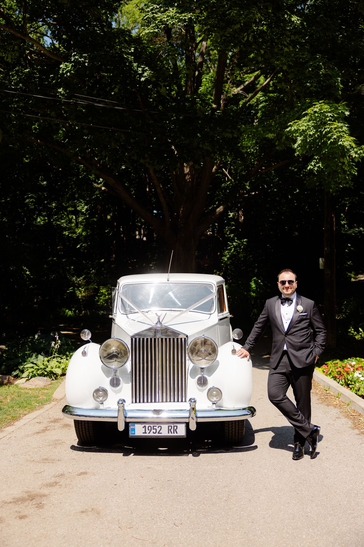 Classic white Rolls-Royce parked on a tree-lined driveway with someone in formal attire standing beside it.