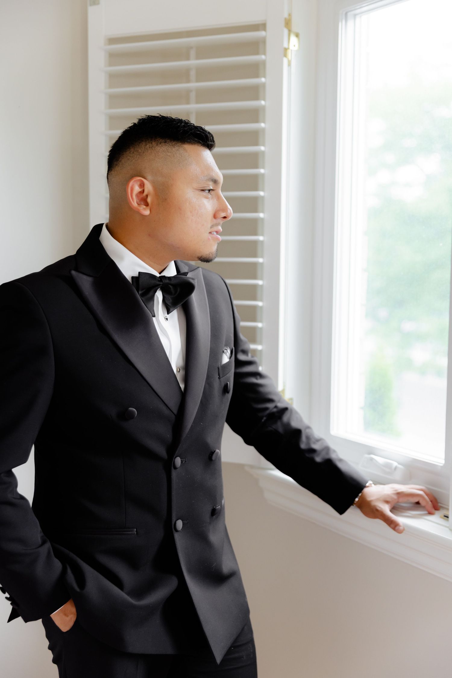 Person in formal black tuxedo gazes thoughtfully through window while standing in bright room with white walls.
